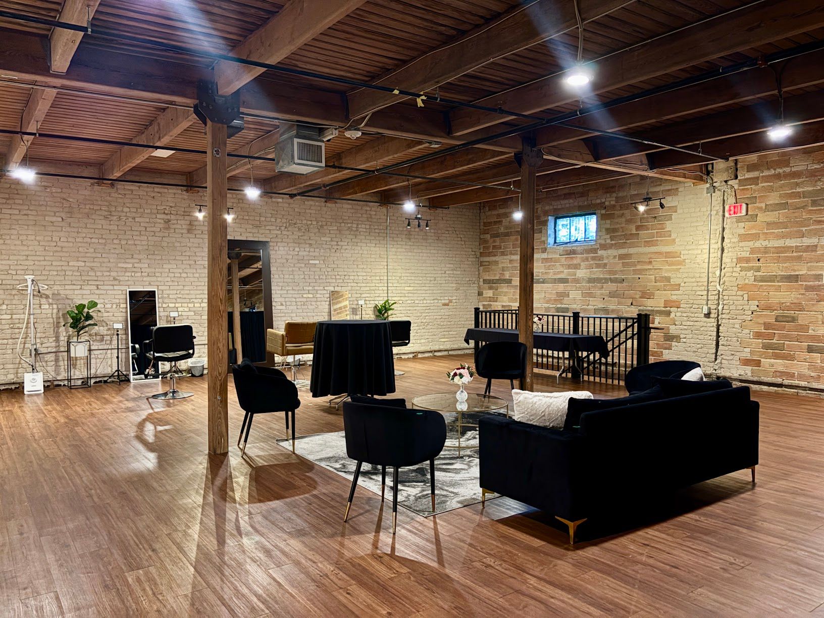 Empty brick-walled event space with wooden floor, black chairs, tables, and lounge seating under exposed beams