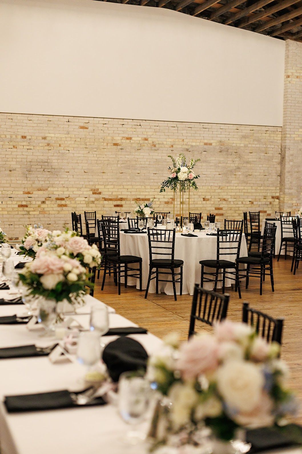 Elegant reception hall with round tables, black chairs, and white floral centerpieces beneath a high wood ceiling