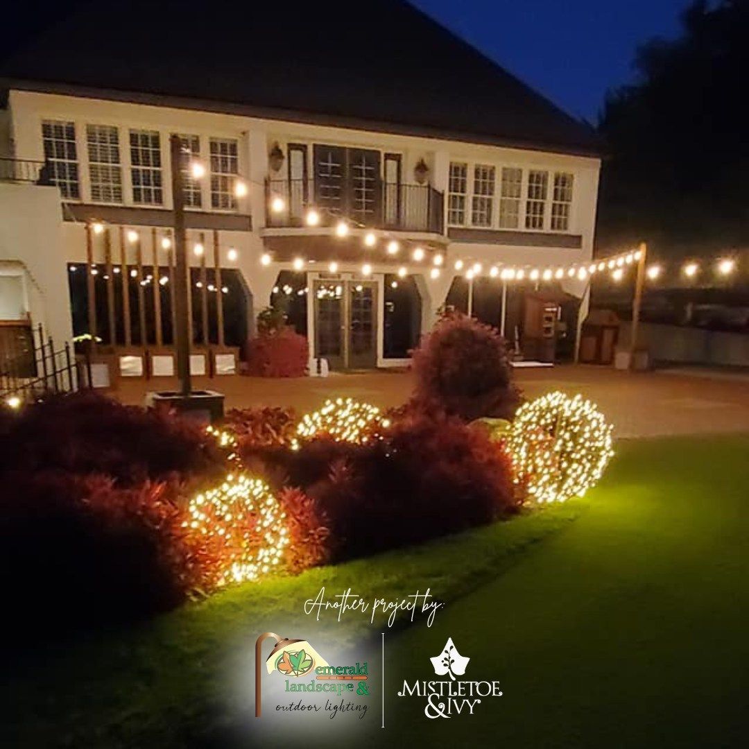 String-lit garden at night beside a house with lit windows and glowing shrubs on a green lawn