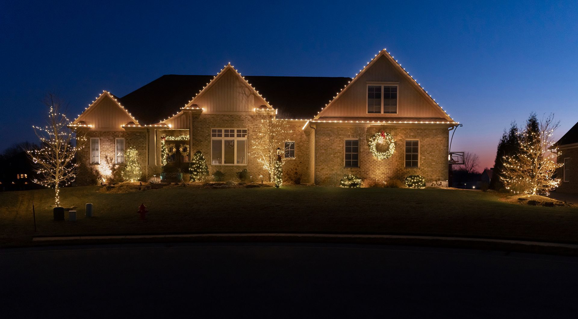 Large stone house lit with warm Christmas lights at dusk, framed by small lit trees.