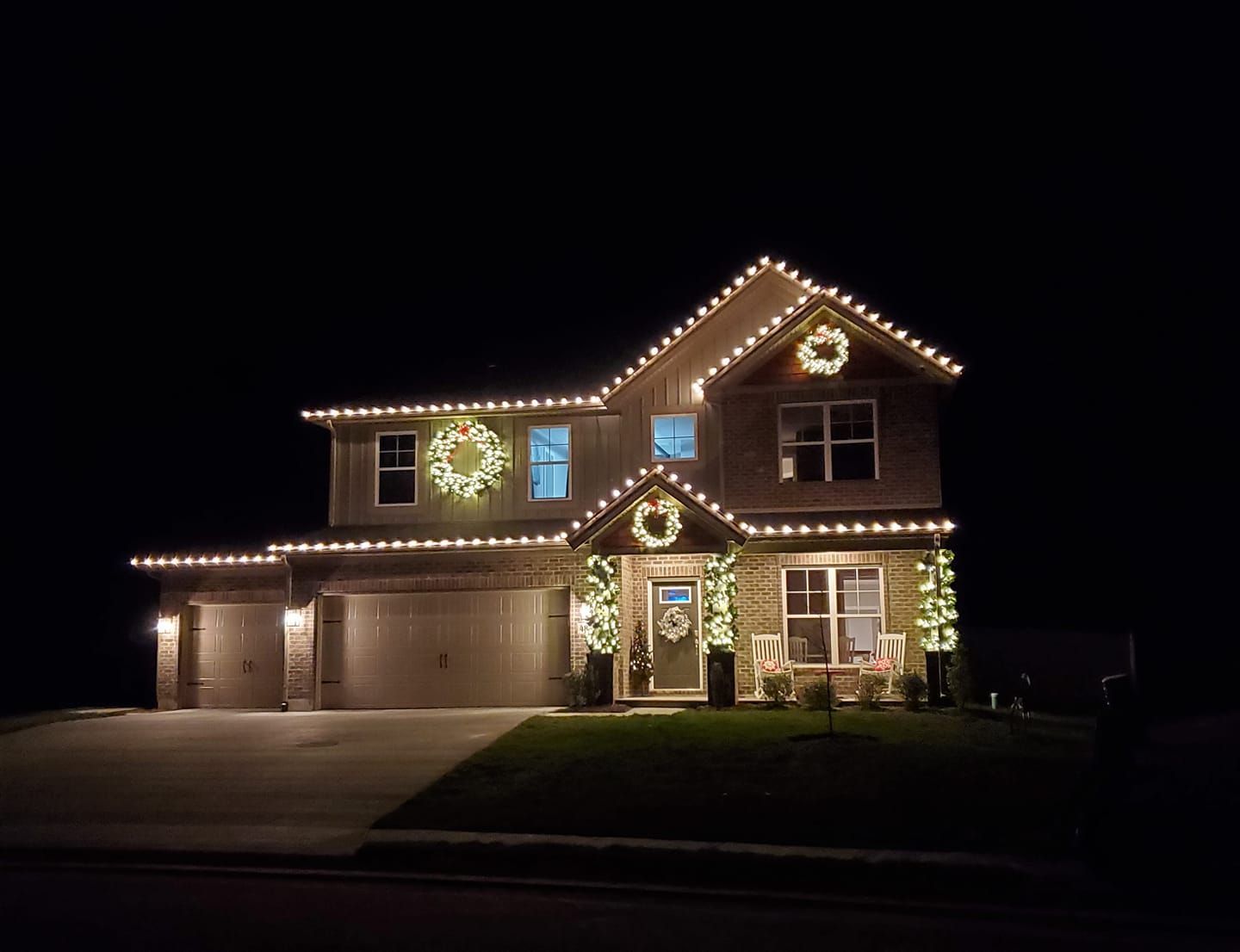 Two-story house at night outlined with warm white holiday lights, with a lit garage and front entry.
