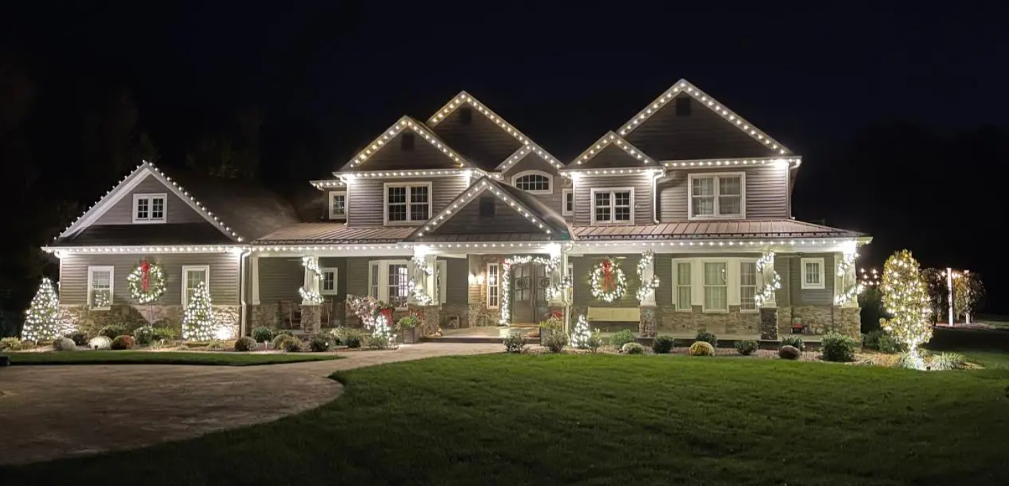 Large house at night decorated with white holiday lights and wreaths, lit lawn and driveway