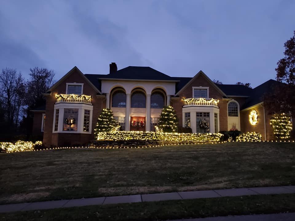 Large house at dusk outlined with warm holiday lights along the roof and shrubs