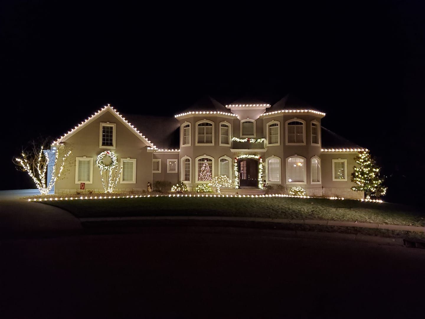 House outlined with warm white Christmas lights at night, with a dark yard and black sky.