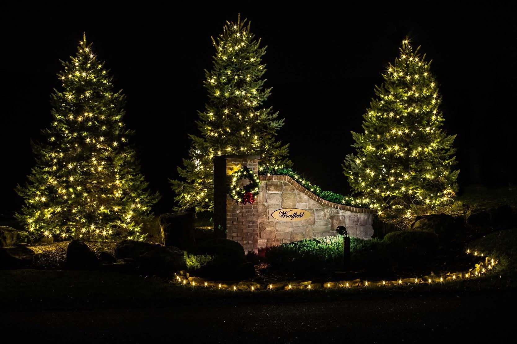 Three lit Christmas trees glowing around a stone wall and house at night
