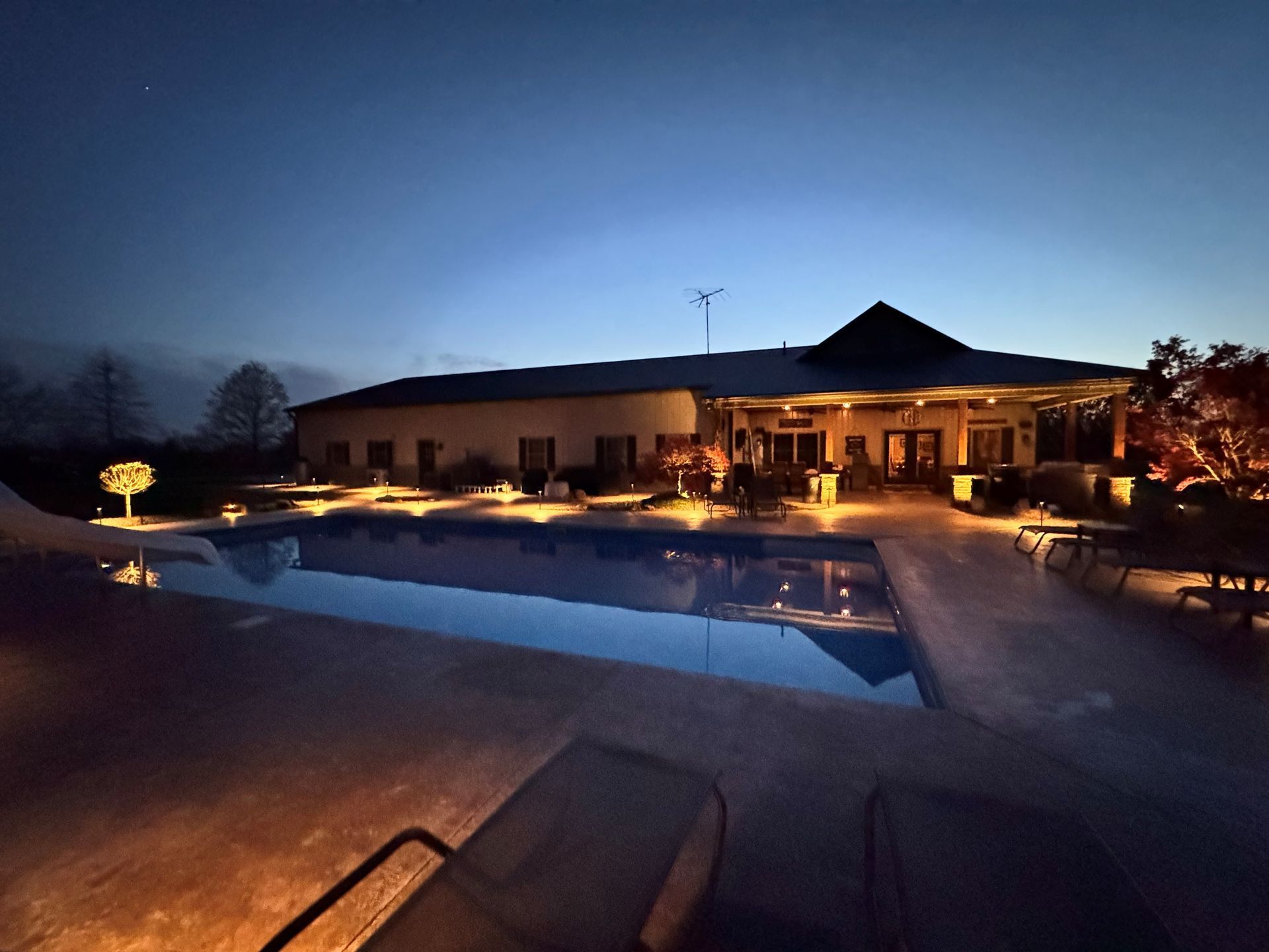 Lit poolside patio at dusk with a house and glowing lights reflecting on the water