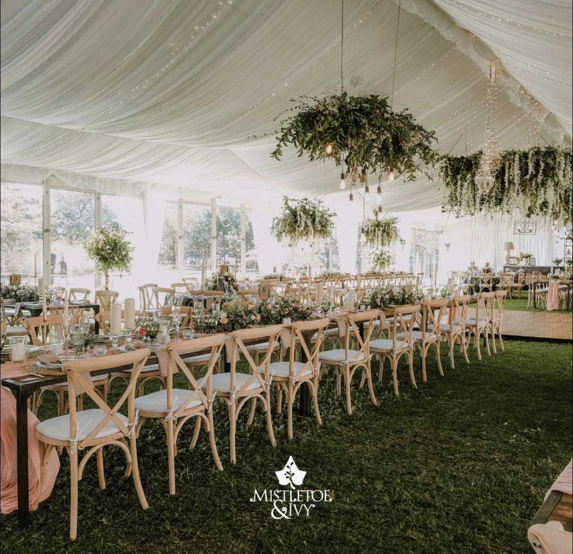 Long wooden tables set for a wedding reception in a white tent on grass.