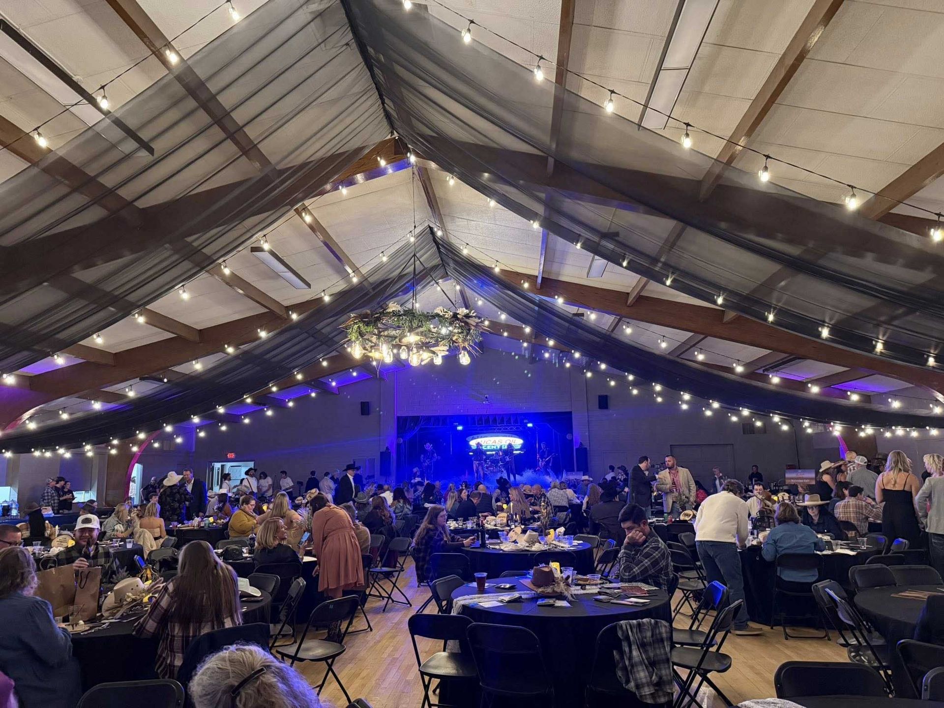 Event in a large hall with tables, people, and a stage with blue lighting. String lights are hung.