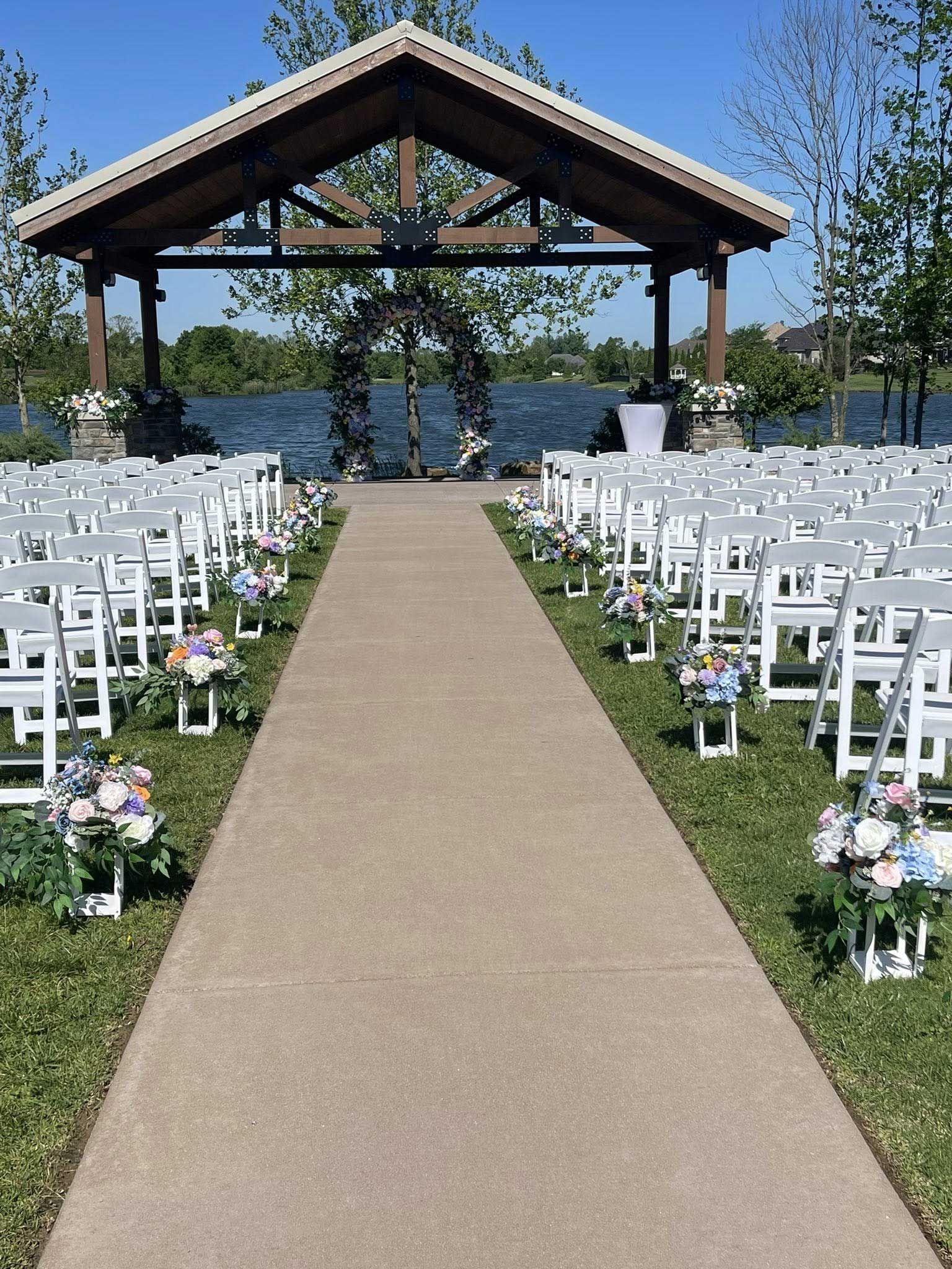 Wedding ceremony setup with white chairs, floral aisle decorations, and gazebo overlooking a lake.