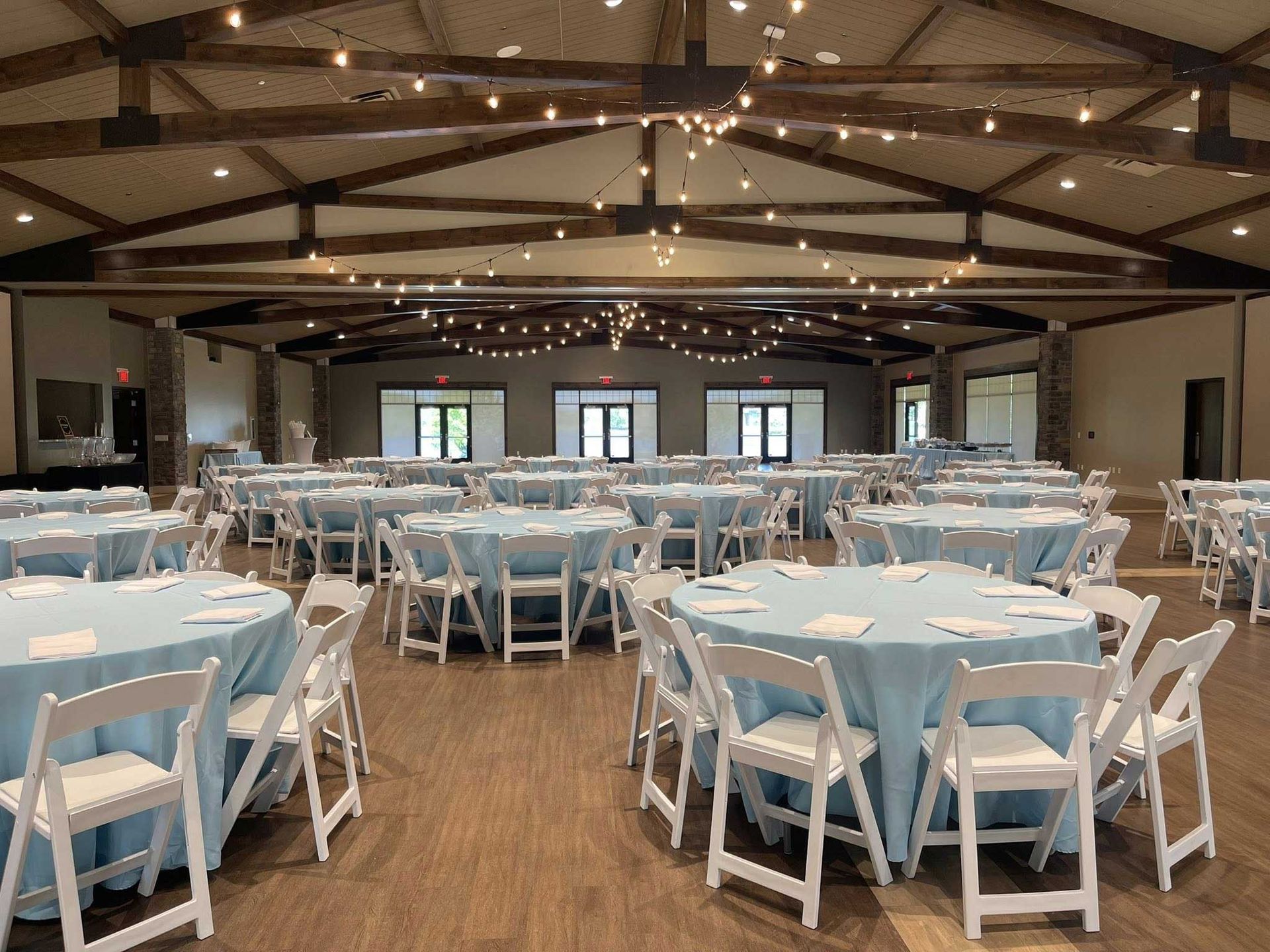 Large event space with round tables covered in light blue cloths, white chairs, and string lights.