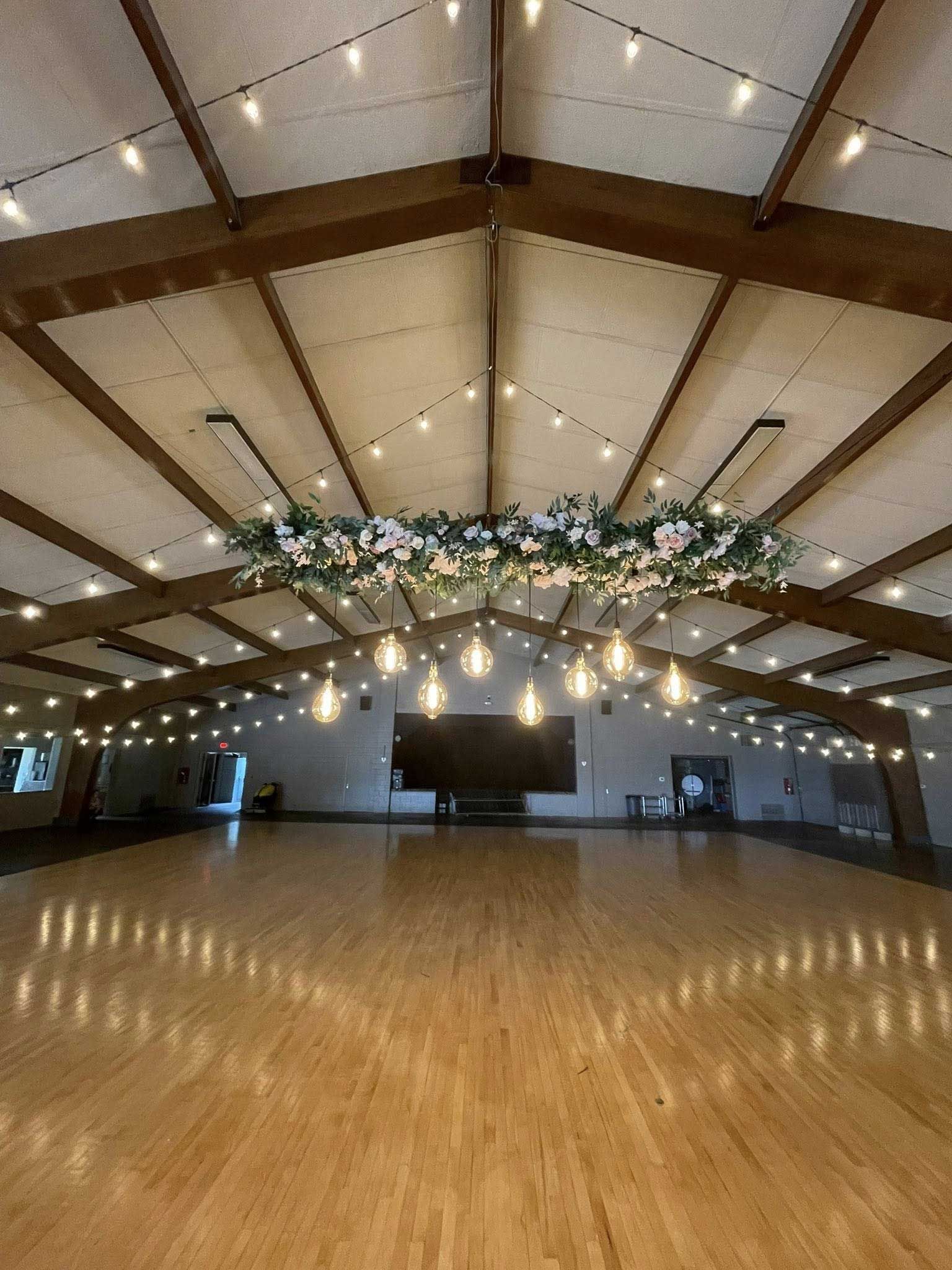 Ballroom with wood floor, high ceiling, floral chandelier, and string lights.