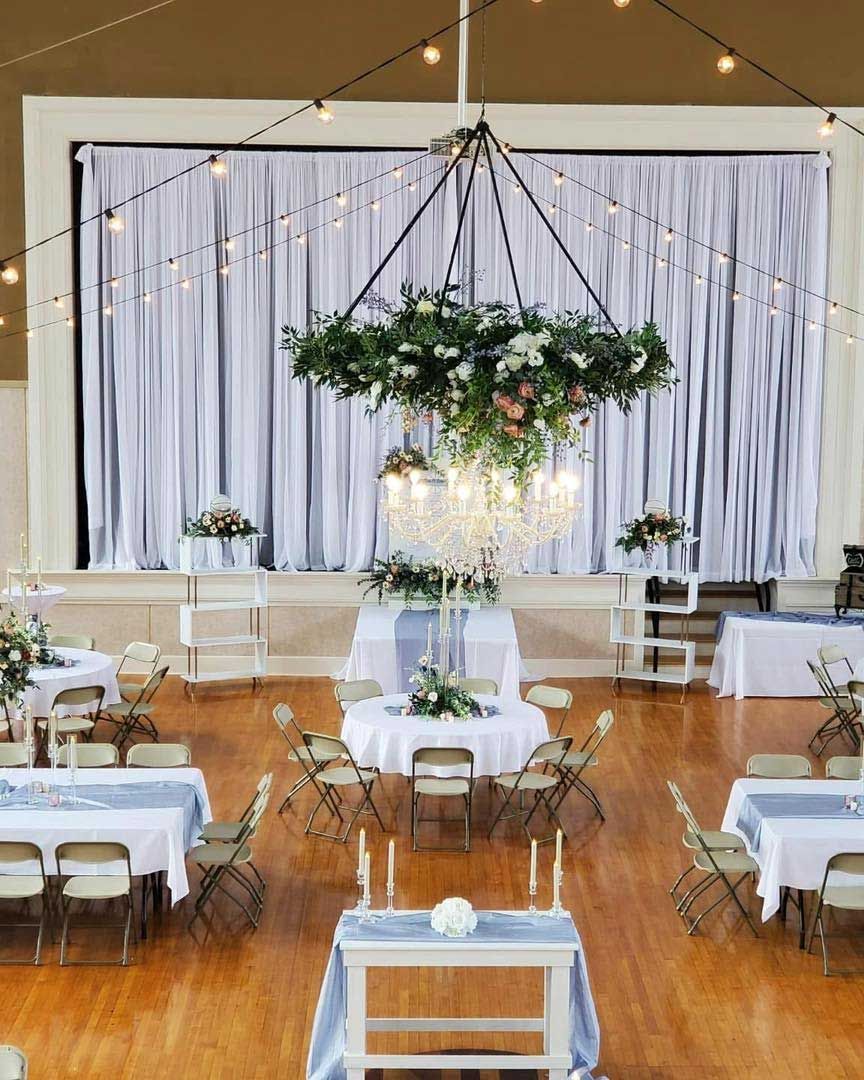 Wedding reception hall with floral chandelier, draped backdrop, tables set for guests, and string lights.