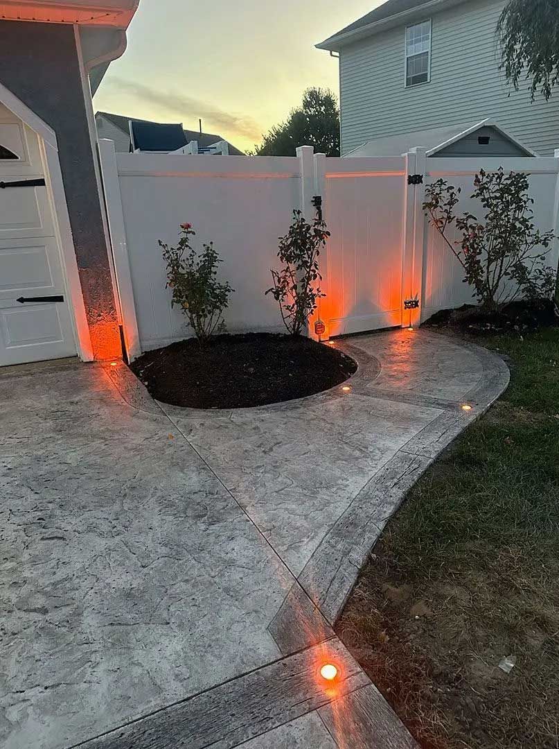 Concrete patio with orange spotlights, a white fence, and small shrubs at dusk.