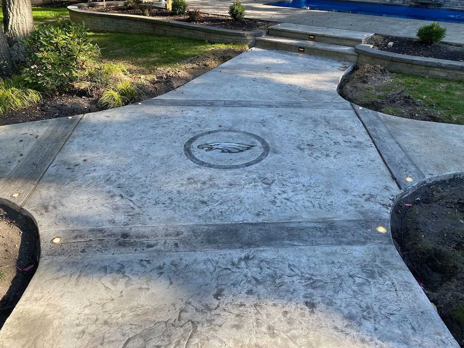 Concrete walkway with a circular decorative inlay and built-in lights, edged by greenery.