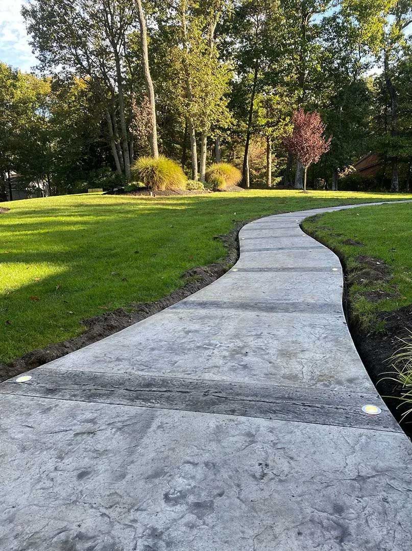 Curved concrete path through a green lawn, leading to trees in the background.