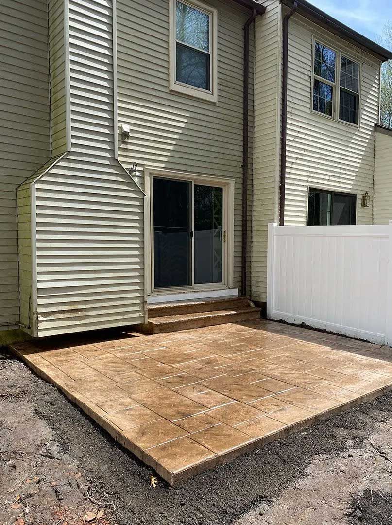 A tan concrete patio outside a beige house with a sliding glass door and white fence.
