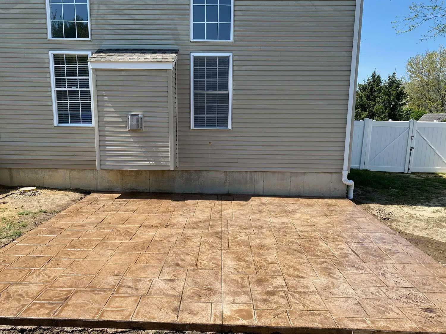 Brown stamped concrete patio outside a beige house with several windows and a white fence.