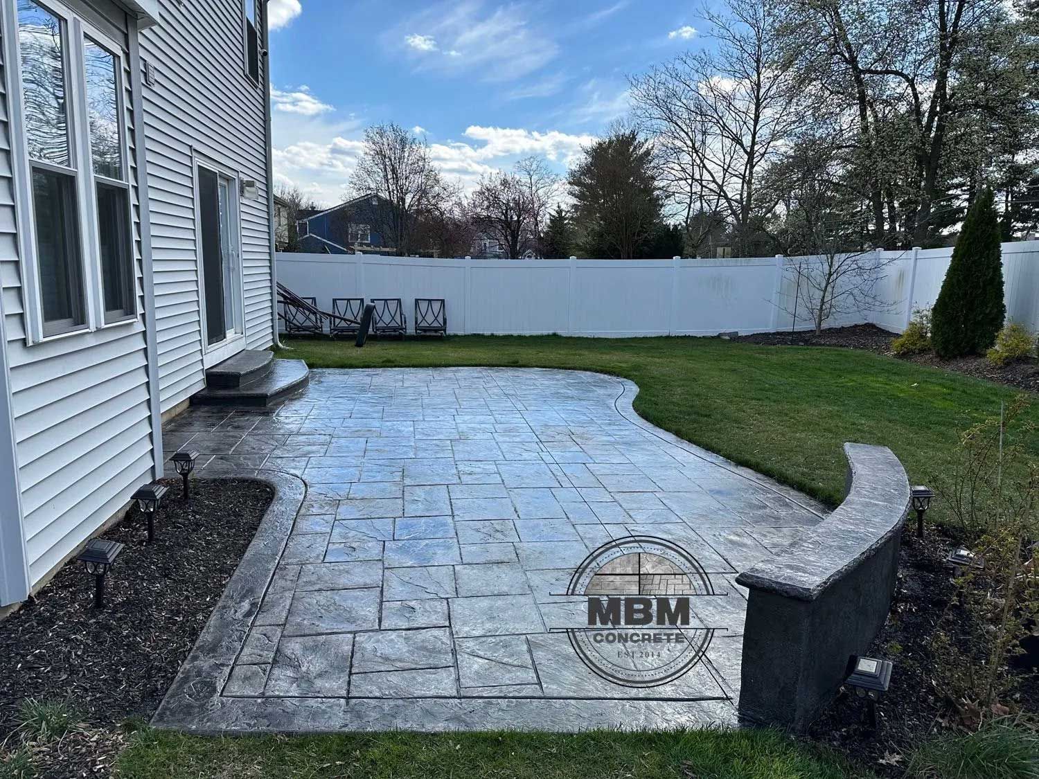 Concrete patio next to a house, bordered by grass, with a low retaining wall.