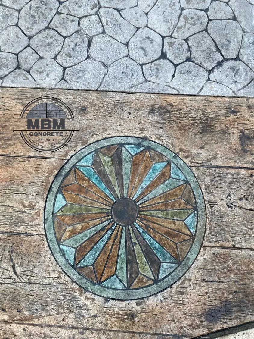 Decorative concrete patio with a circular, geometric design in earth tones. MBM Concrete logo visible.