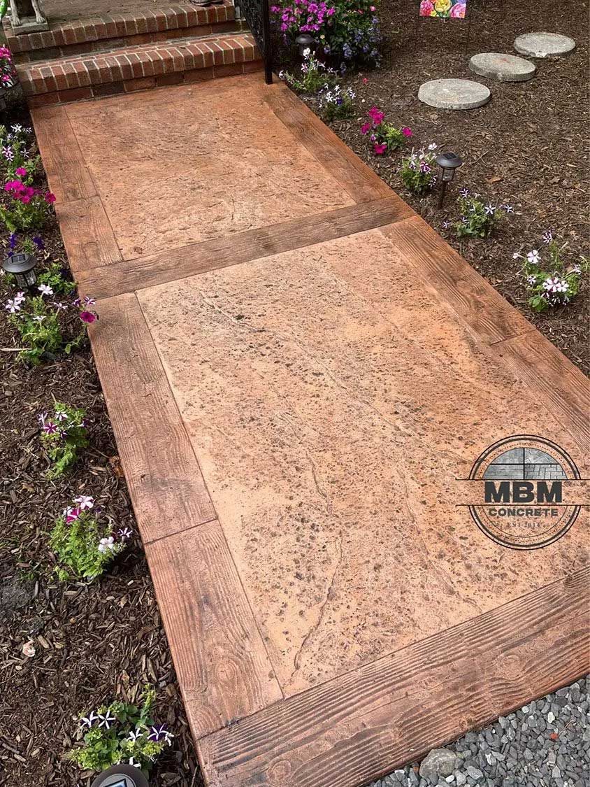 Stained concrete walkway with decorative border, leading to brick steps.  Flowers and landscaping on either side.