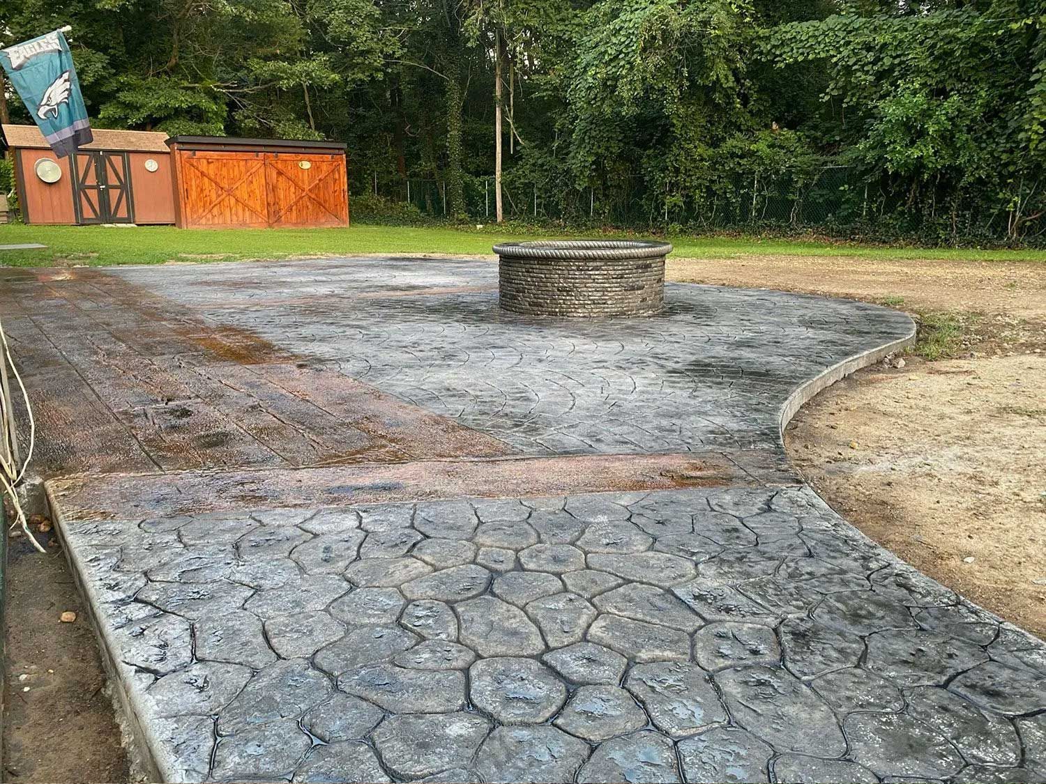 Concrete patio with fire pit, leading to a wooden shed in a grassy outdoor setting.