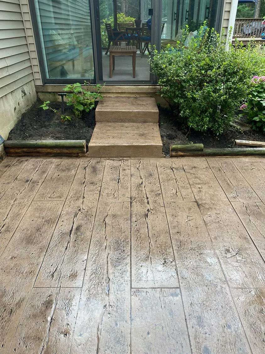 Concrete patio with textured pattern leading to steps and a glass door. Plants on each side.