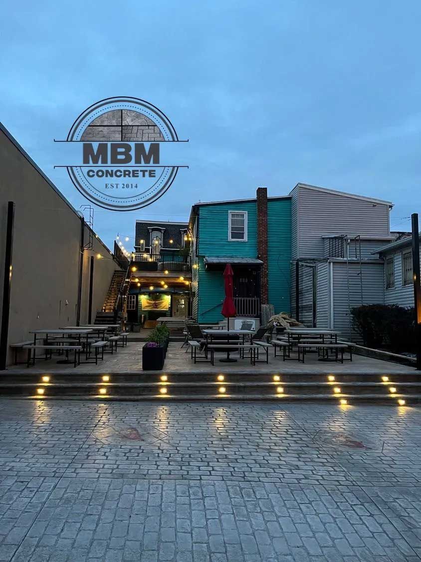 Outdoor patio with tables and seating. Teal building in the background, stamped concrete, evening lighting.
