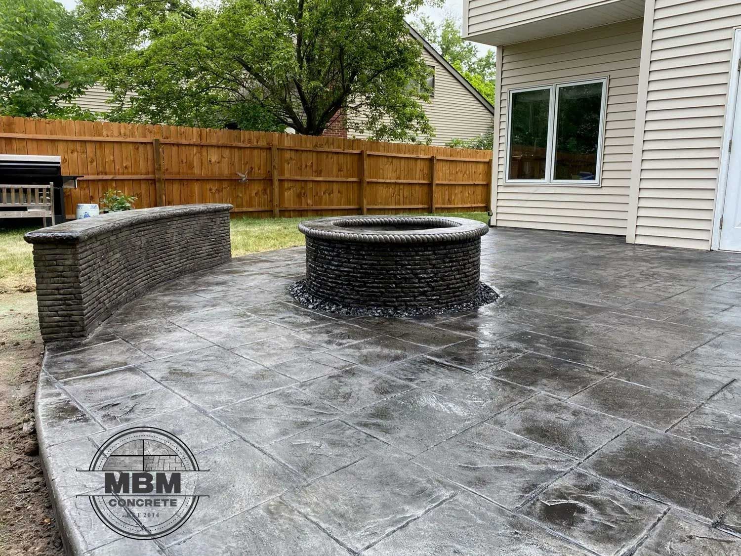 Stamped concrete patio with fire pit and retaining wall, next to a house with a wood fence.