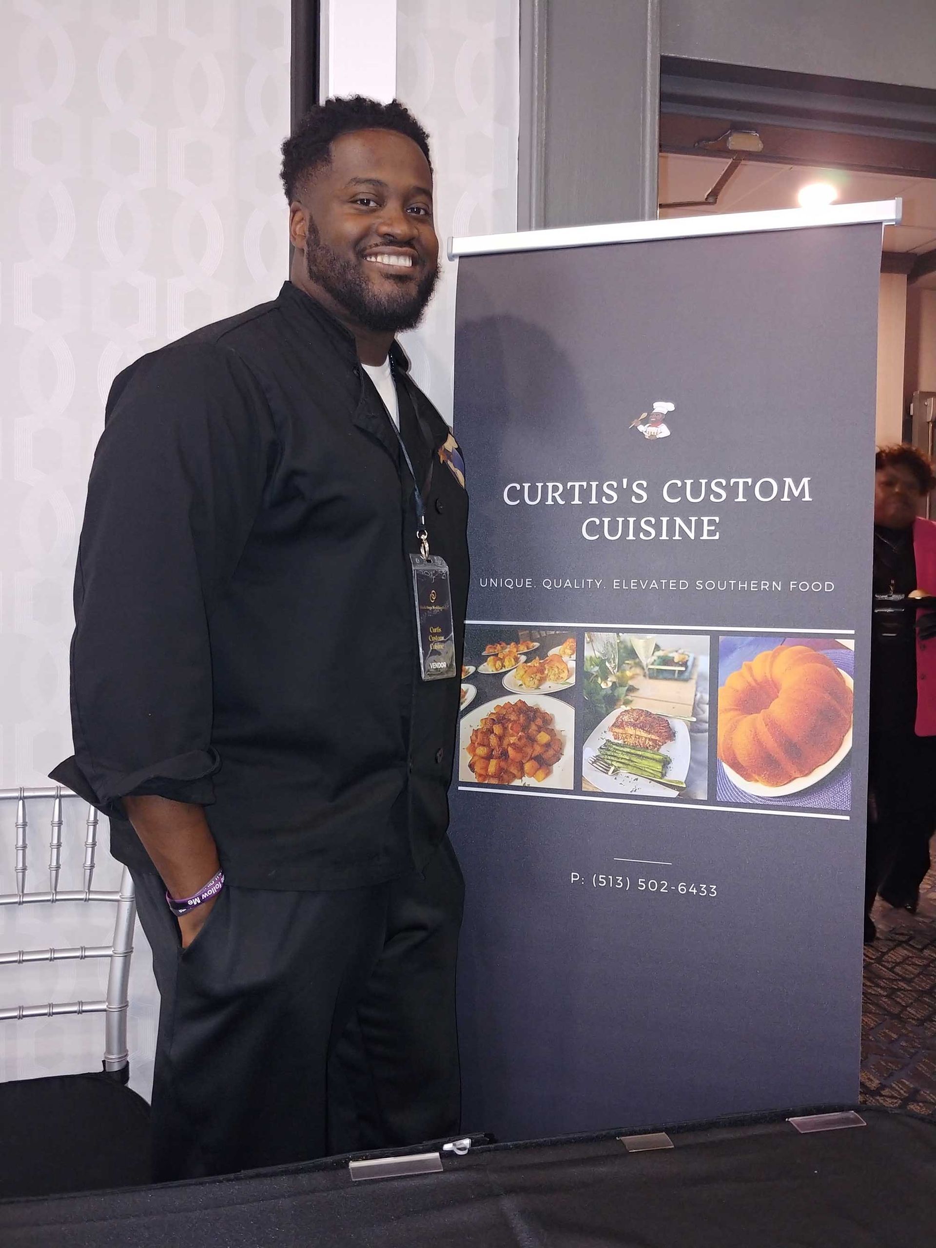 Man in chef's uniform smiles next to banner for 