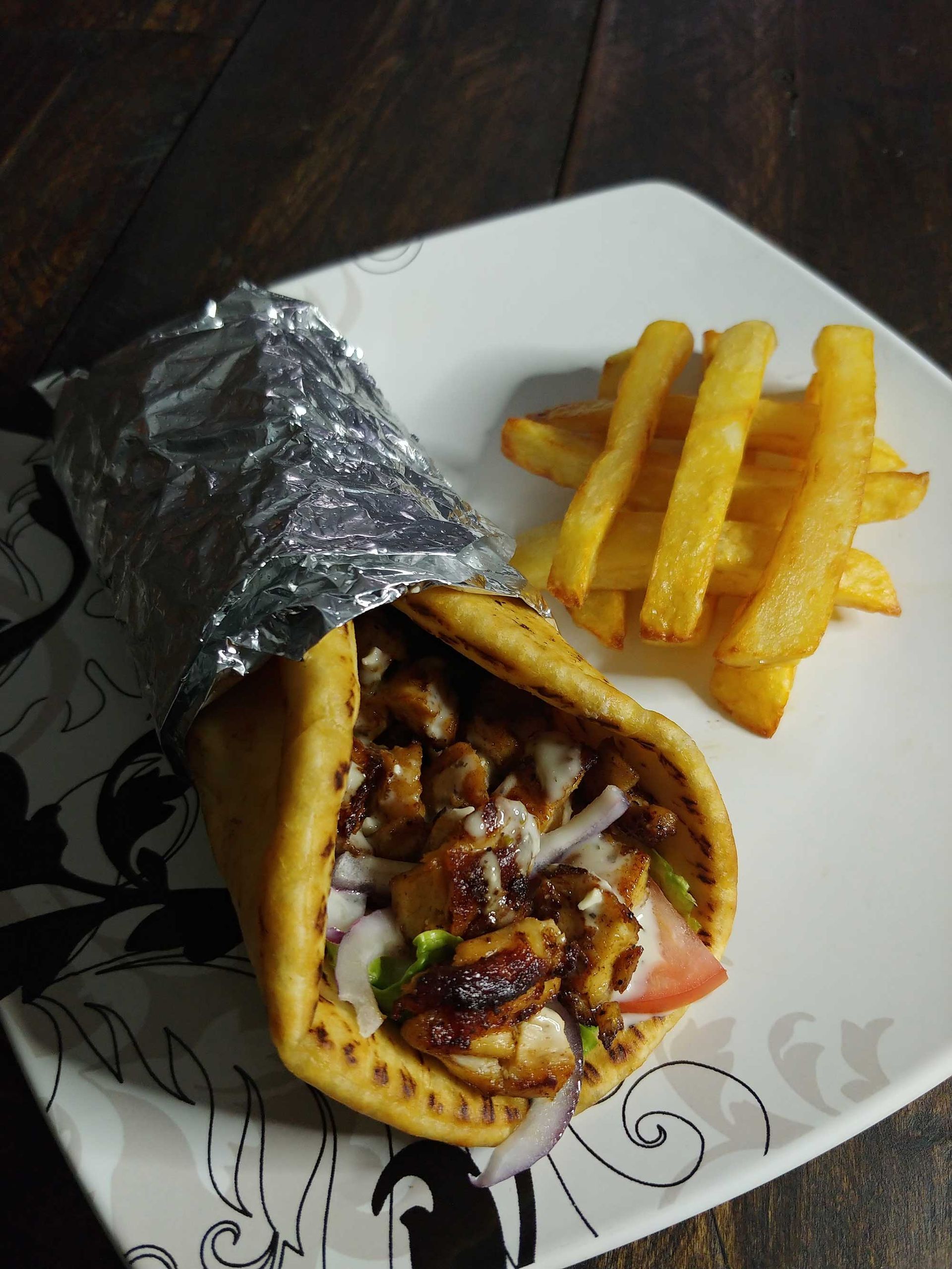 Gyros wrap with fries on a plate. The wrap is wrapped in foil. The fries are golden.