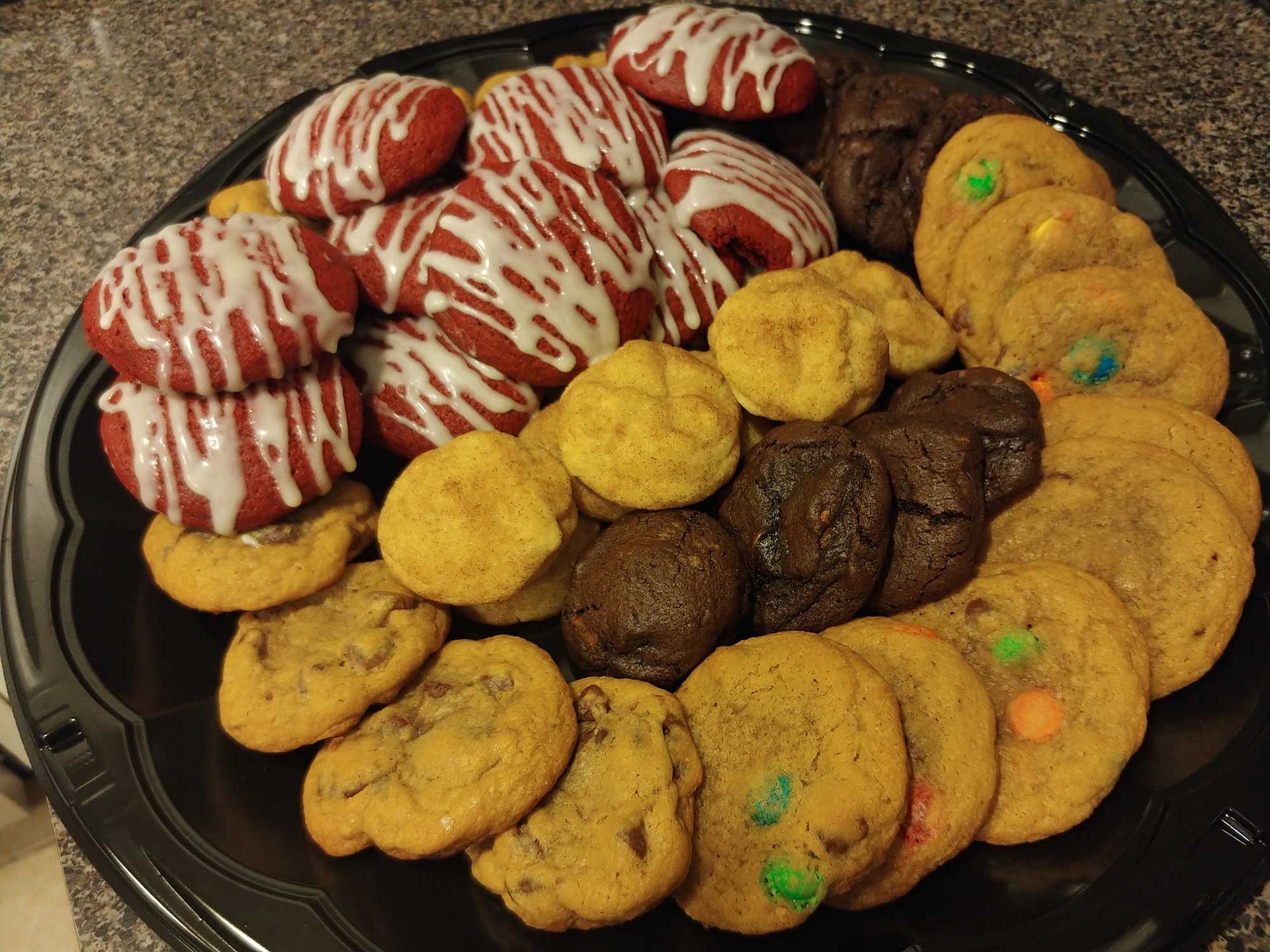 Tray of assorted cookies: red velvet with white drizzle, yellow, and chocolate cookies with candy pieces.