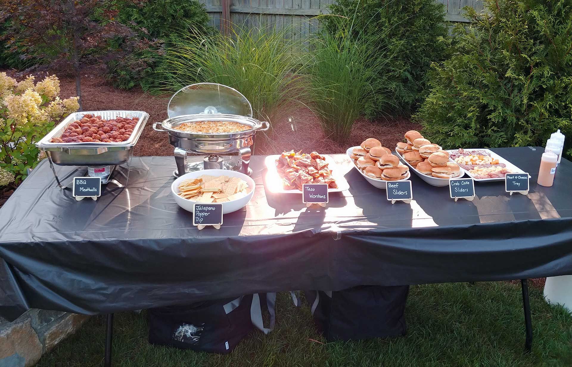Buffet table outdoors with various food dishes: meatballs, mac & cheese, chicken bites, & rolls.