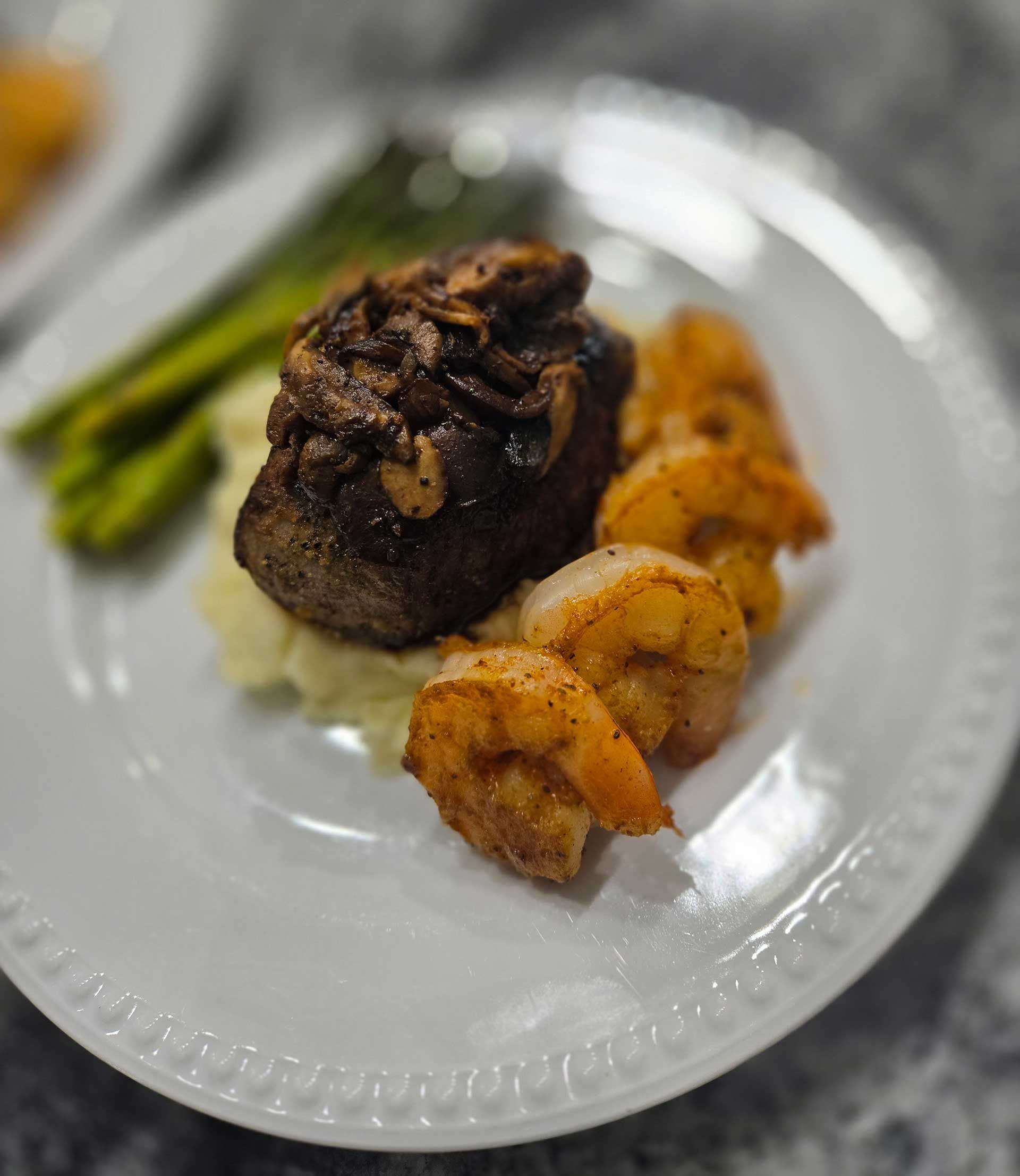 Steak with mushrooms, shrimp, asparagus, and mashed potatoes on a white plate.
