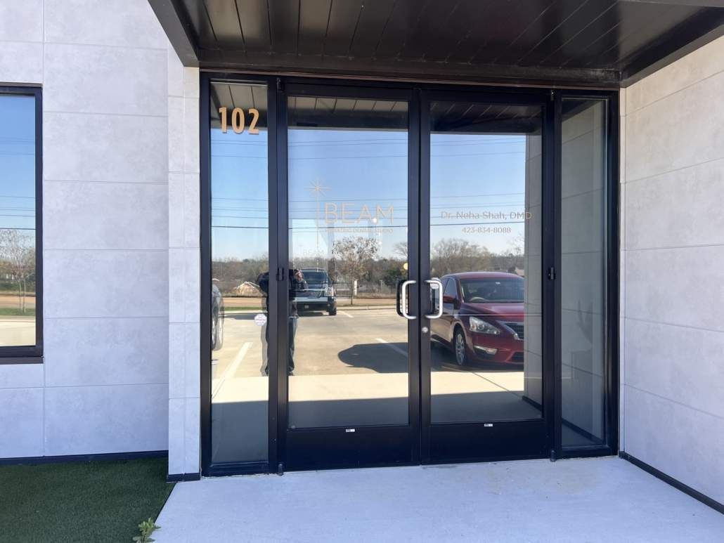 Glass double doors with black frames at the entrance to building 107. Cars and sky reflected in the glass.