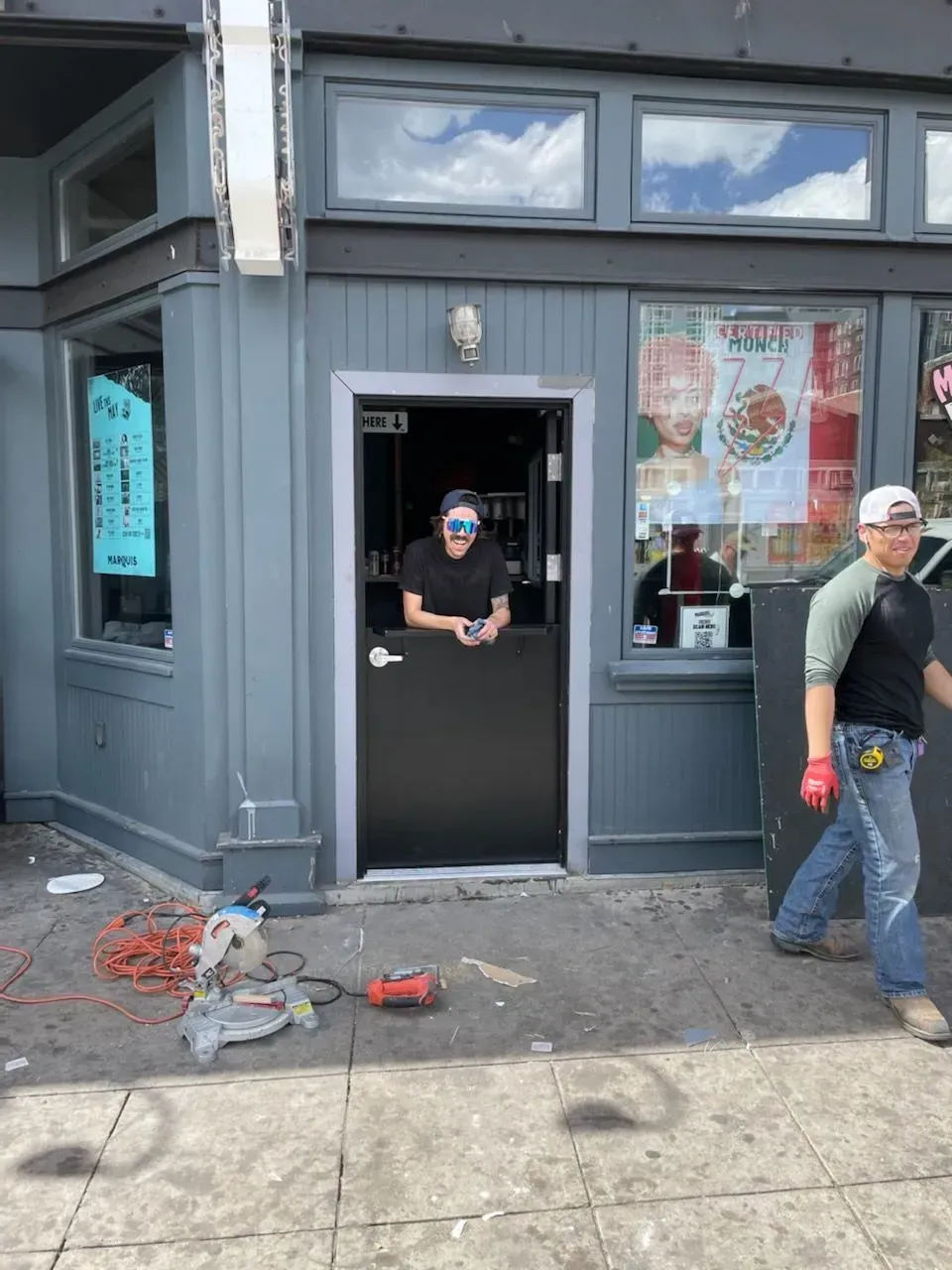 Person stands in doorway of a dark storefront while another person walks past on the sidewalk.