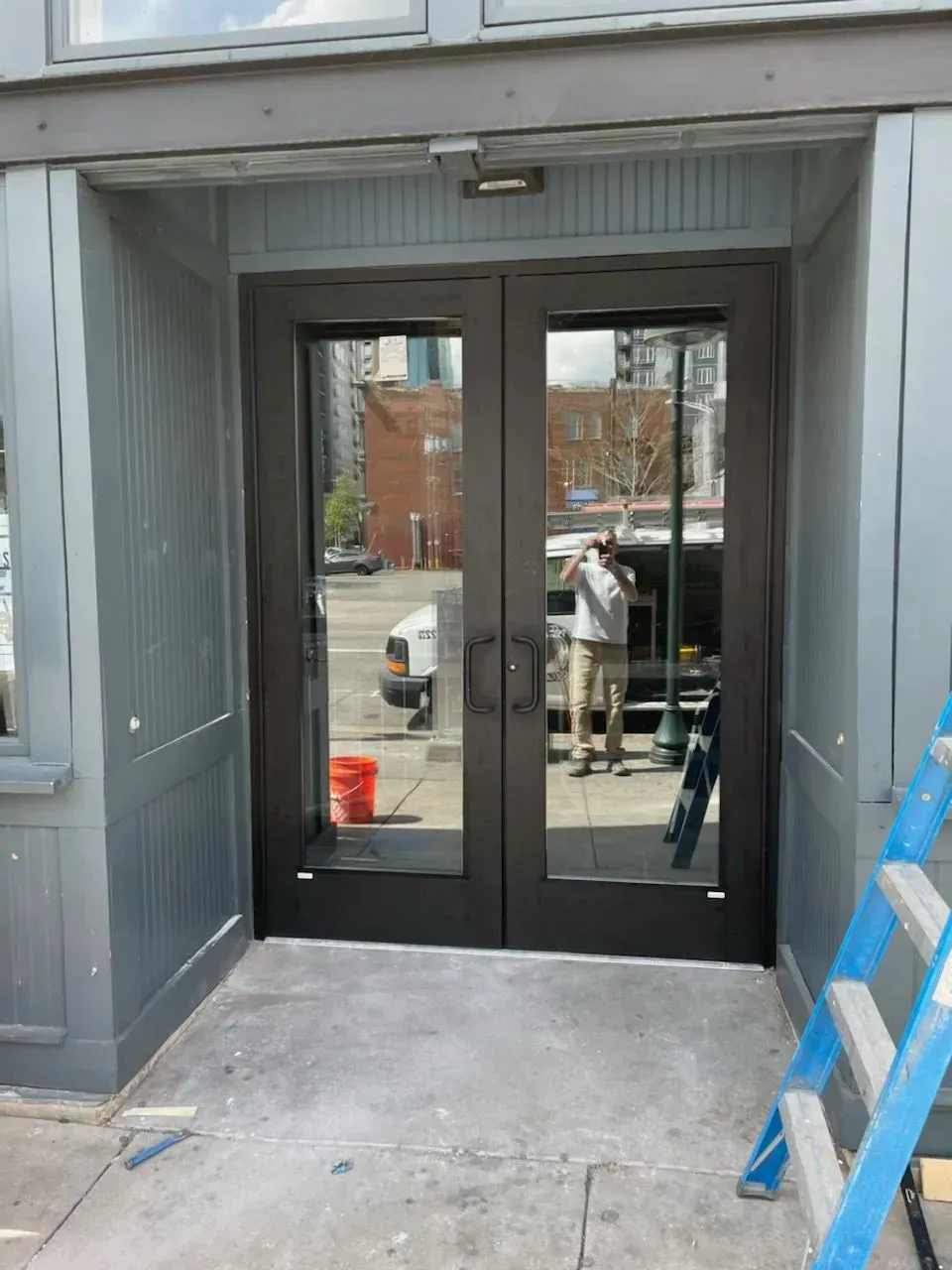 Glass double doors at a building entrance, with a blue ladder on the right and construction tools inside.