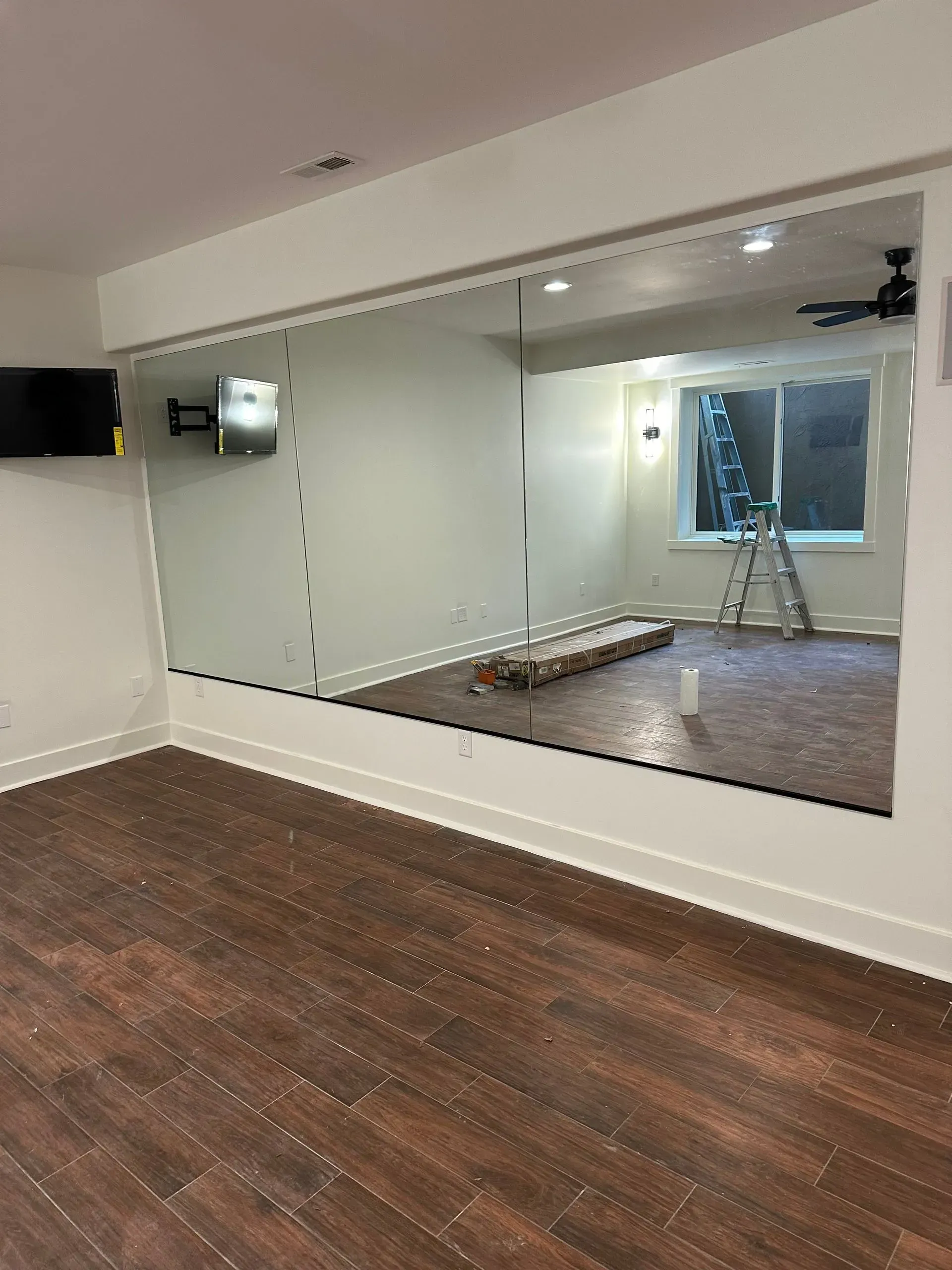 Empty room with dark wood floor, large wall mirror, and a window with a ladder in the reflection.