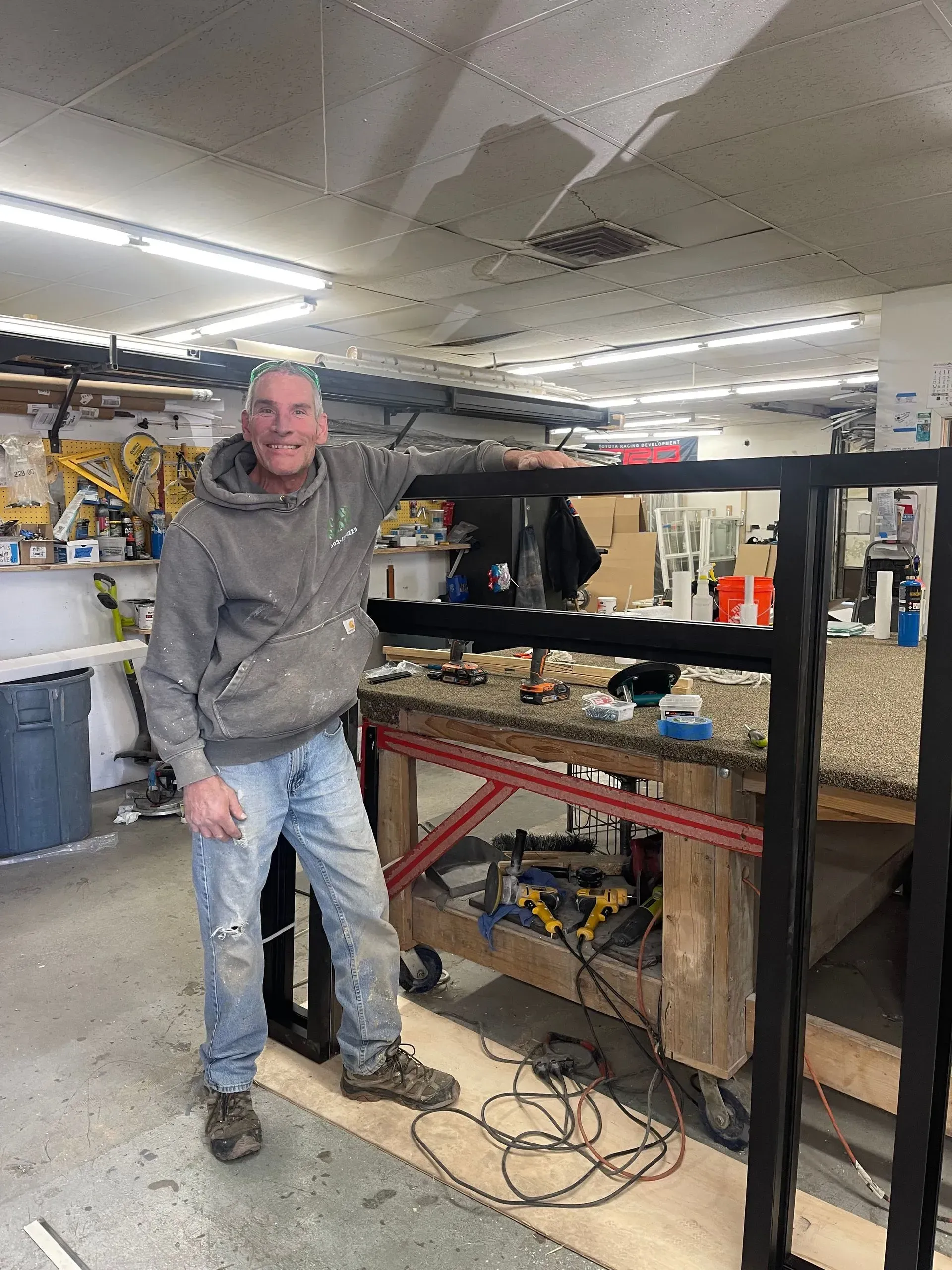 Man standing in a workshop beside a black metal frame, with tools and equipment in the background.