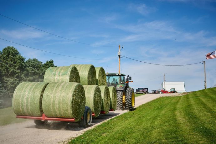 Tractor hauling stacked round hay bales along a rural road under a blue sky