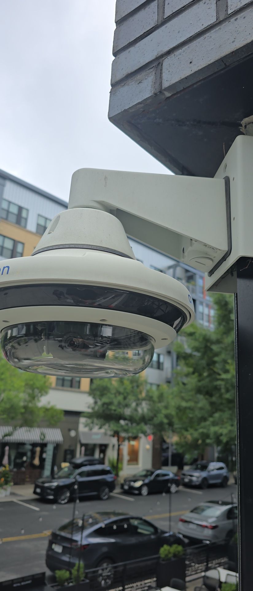 White circular street camera mounted under building awning above parked cars on a city street