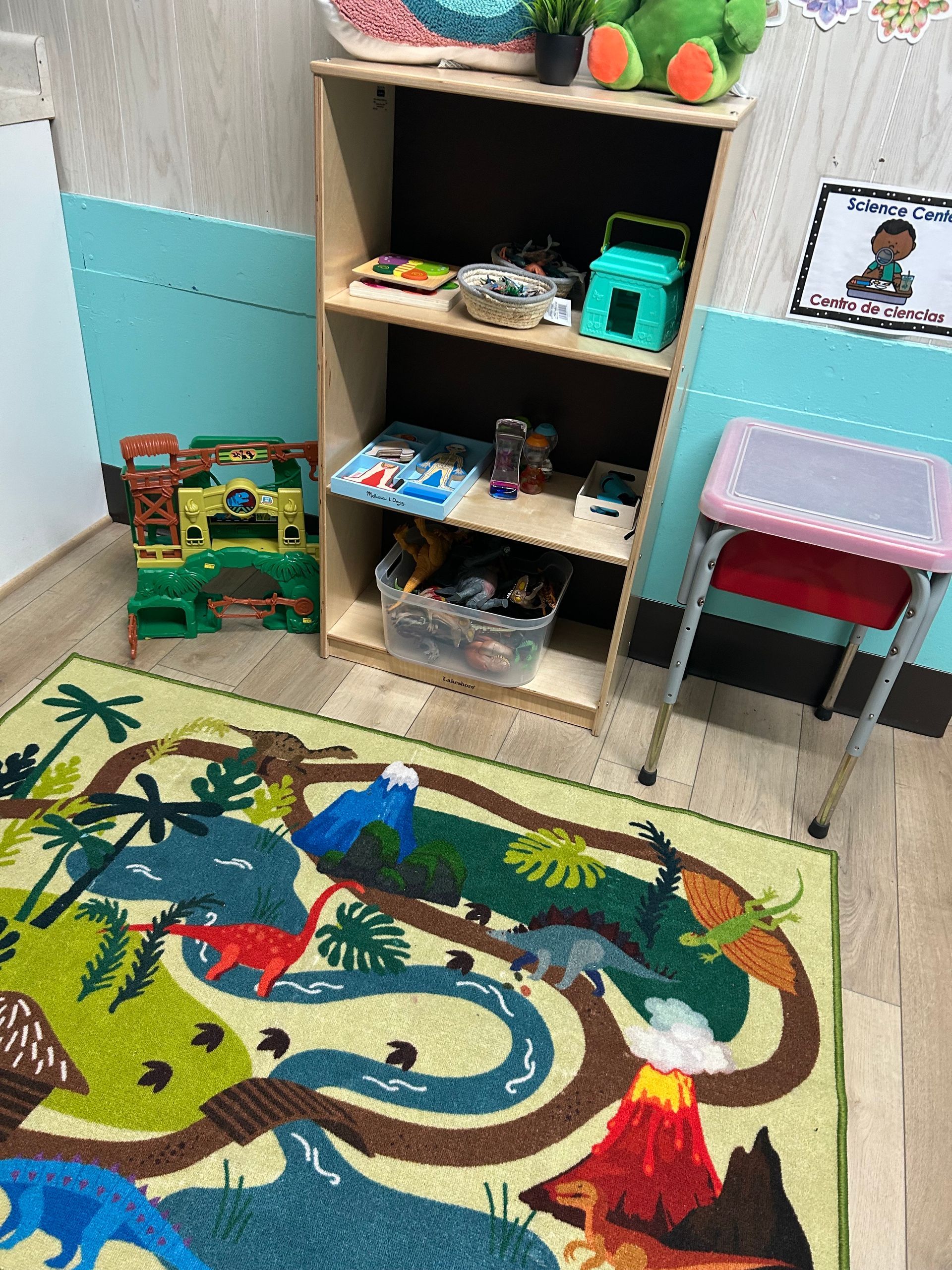 Dinosaur-themed play area with a rug, shelves of toys, and a small stool in a room with blue walls.