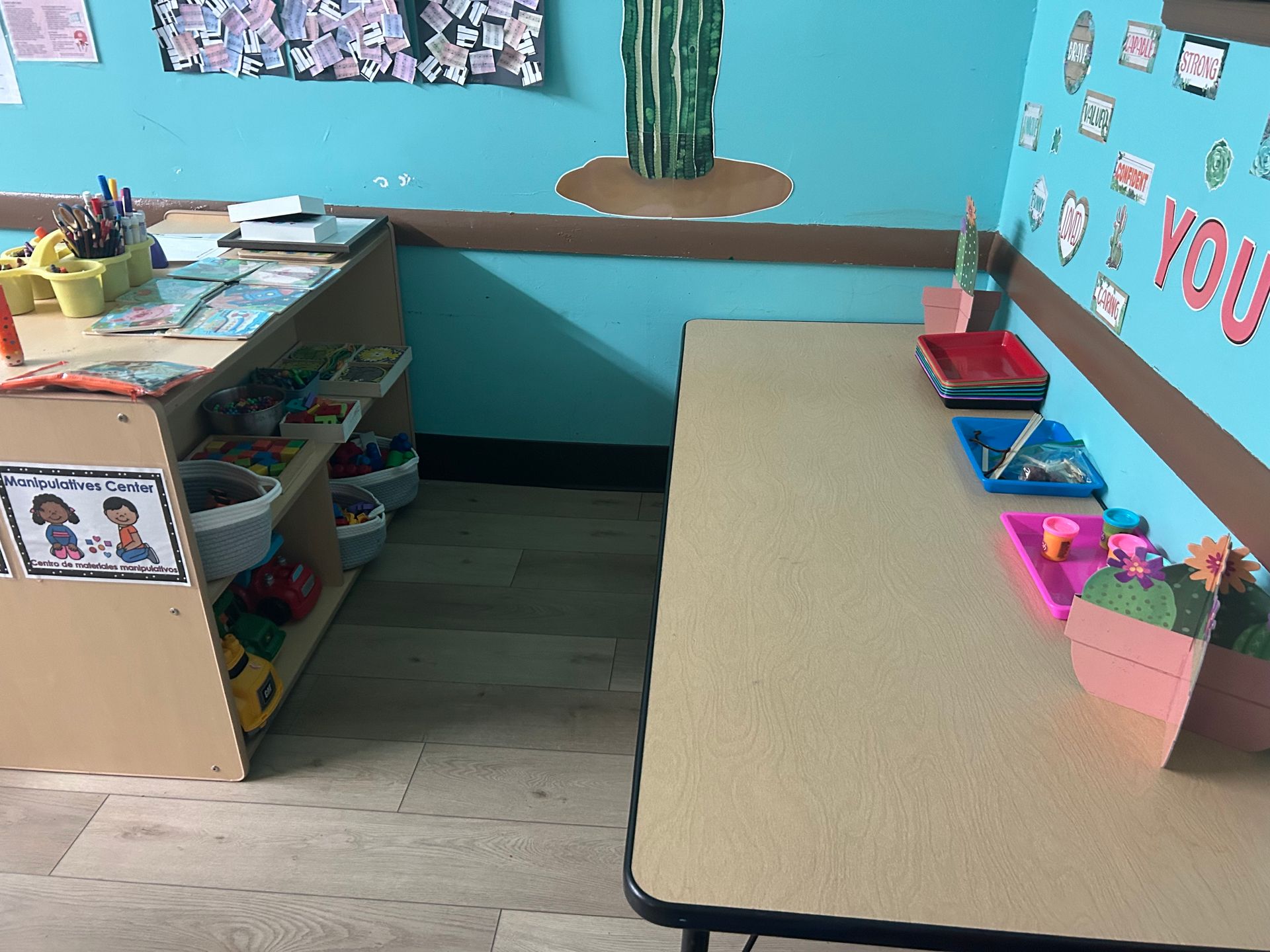 A classroom corner with a table and shelving unit. Crayons, books, and art supplies are present.
