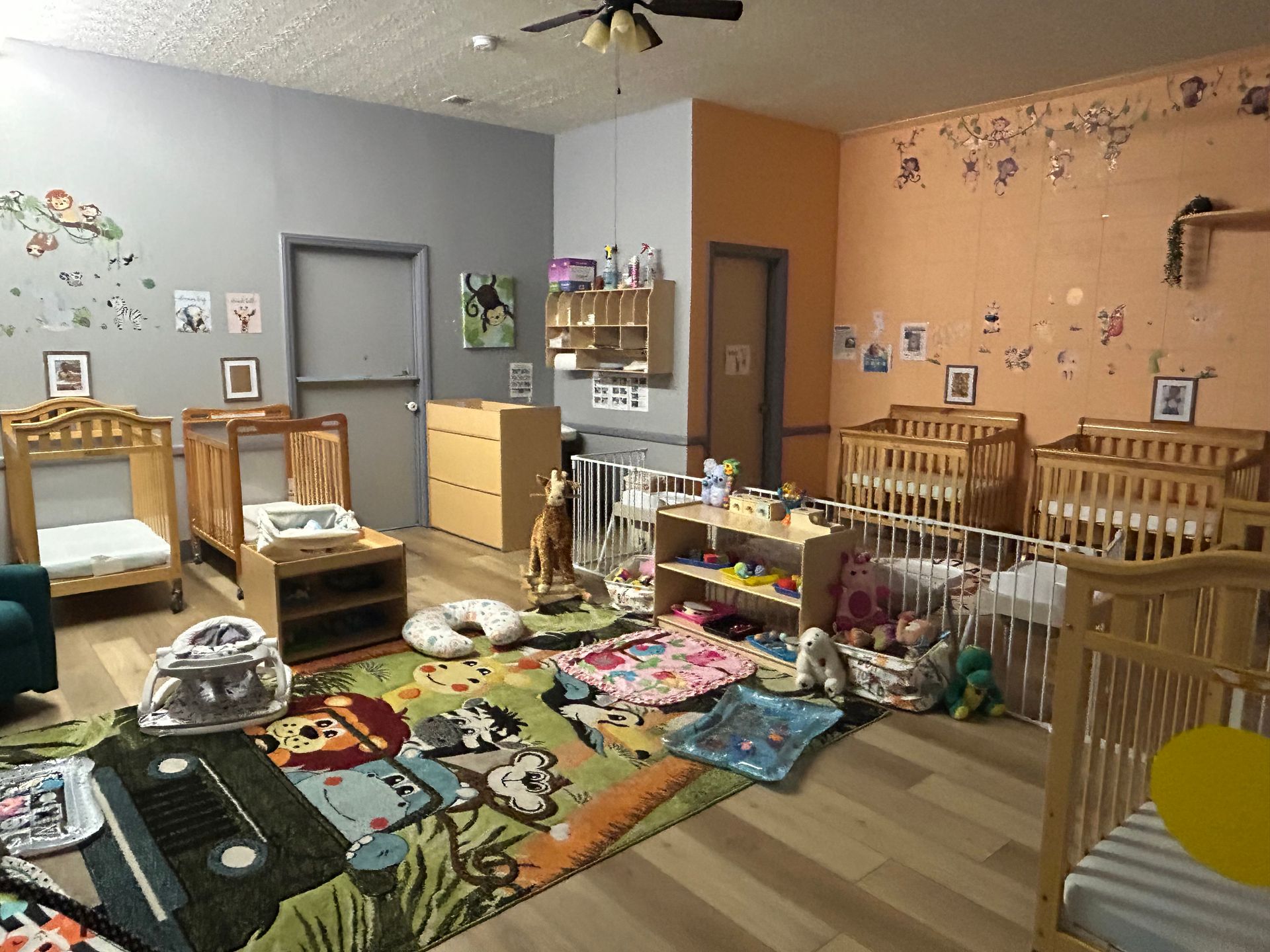 Daycare room with cribs, toys, and colorful wall decorations.