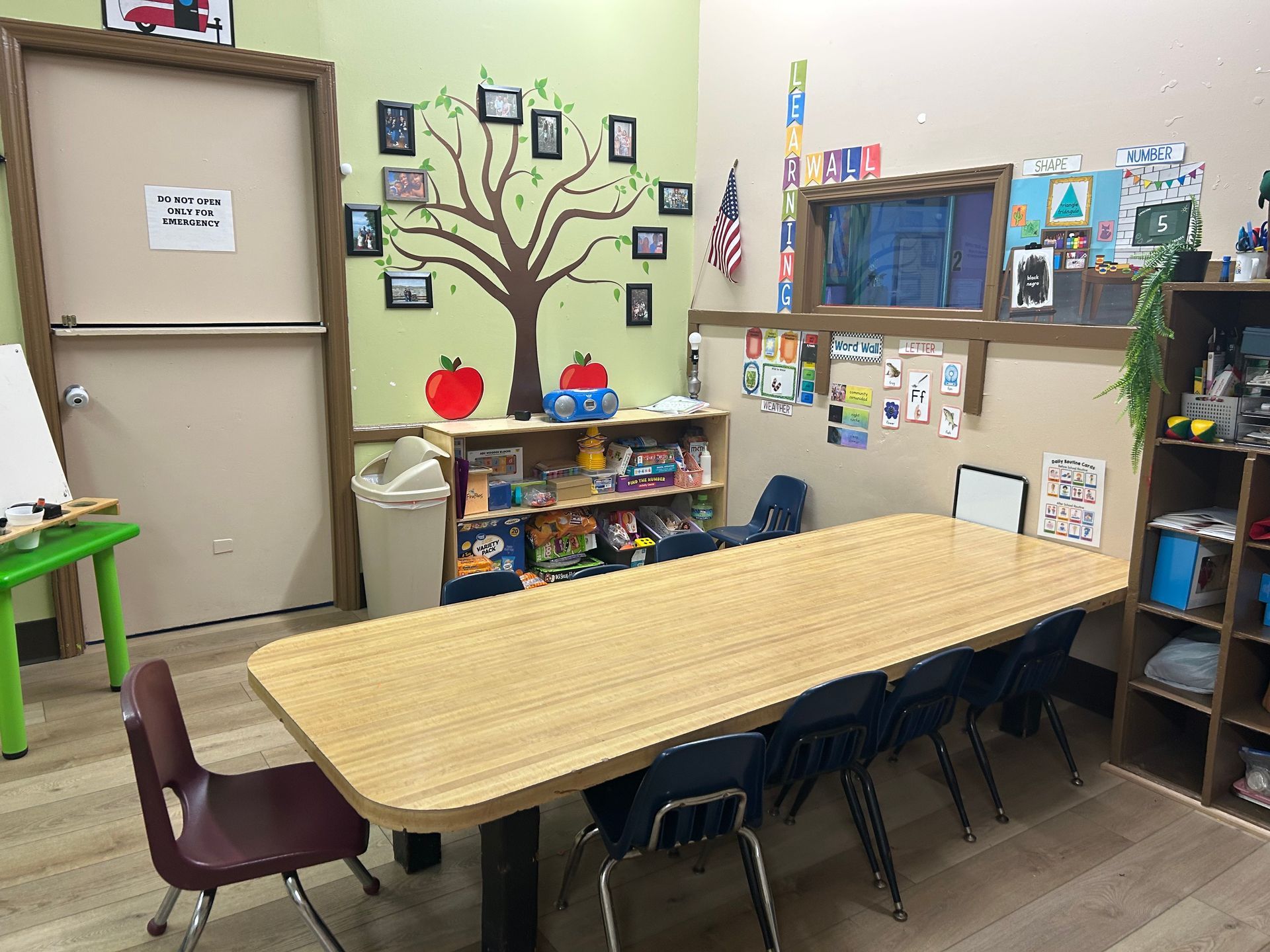 Classroom with a long table and chairs, a tree decal, shelves with toys, and a whiteboard.