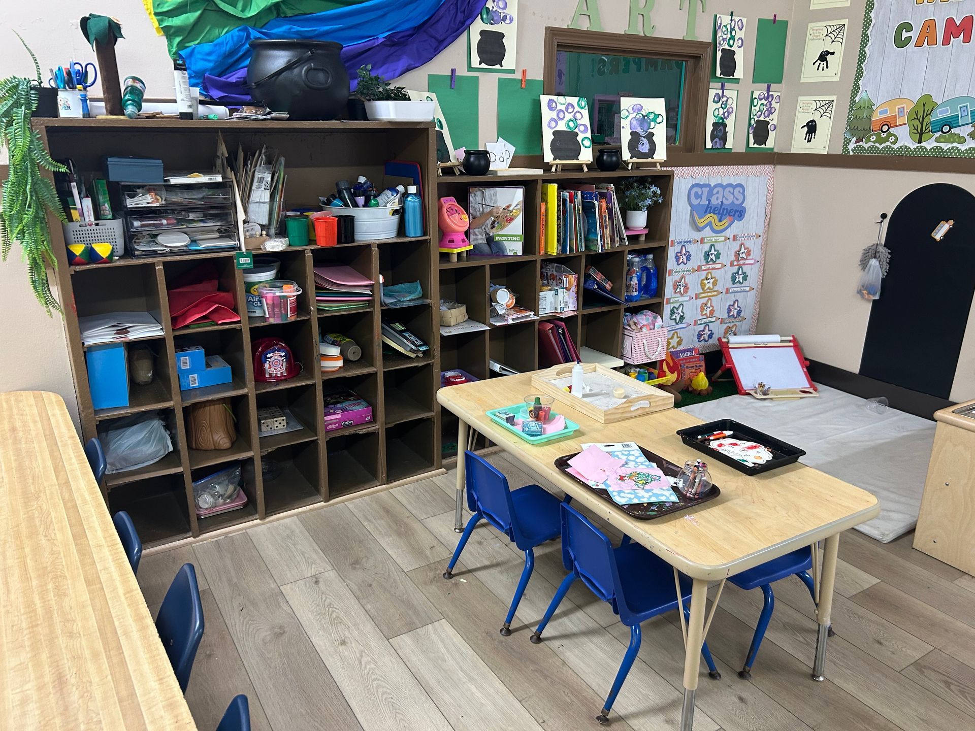 A classroom with storage shelves, tables with trays, and children's artwork is likely a preschool or daycare setting.