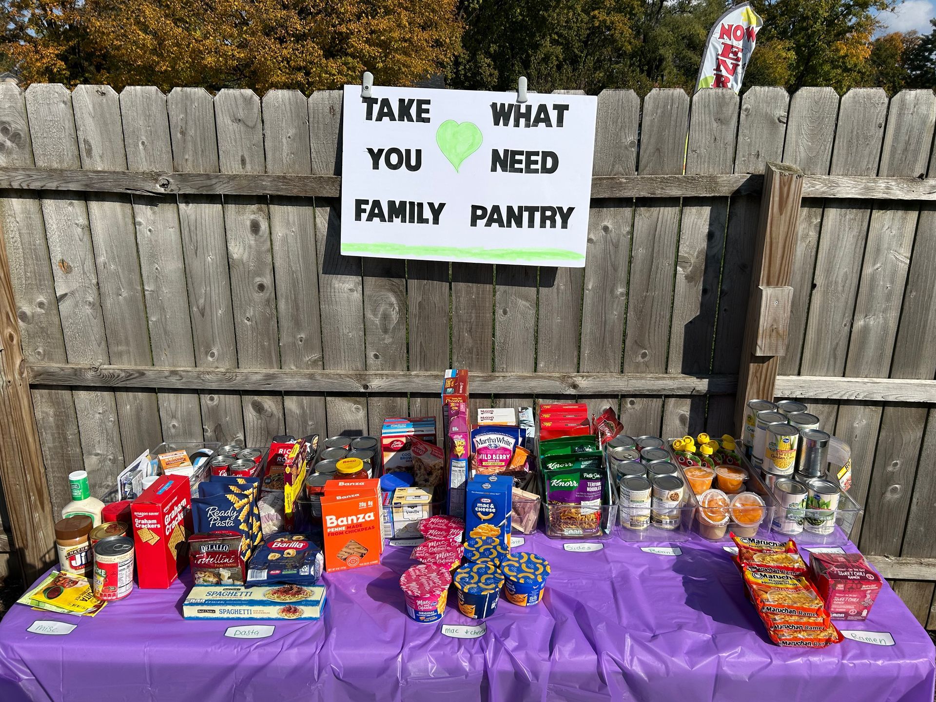 A family pantry, a purple-covered table with food items in front of a wooden fence, sign reads: 
