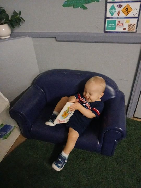 A toddler sits in a blue armchair, holding a book, smiling
