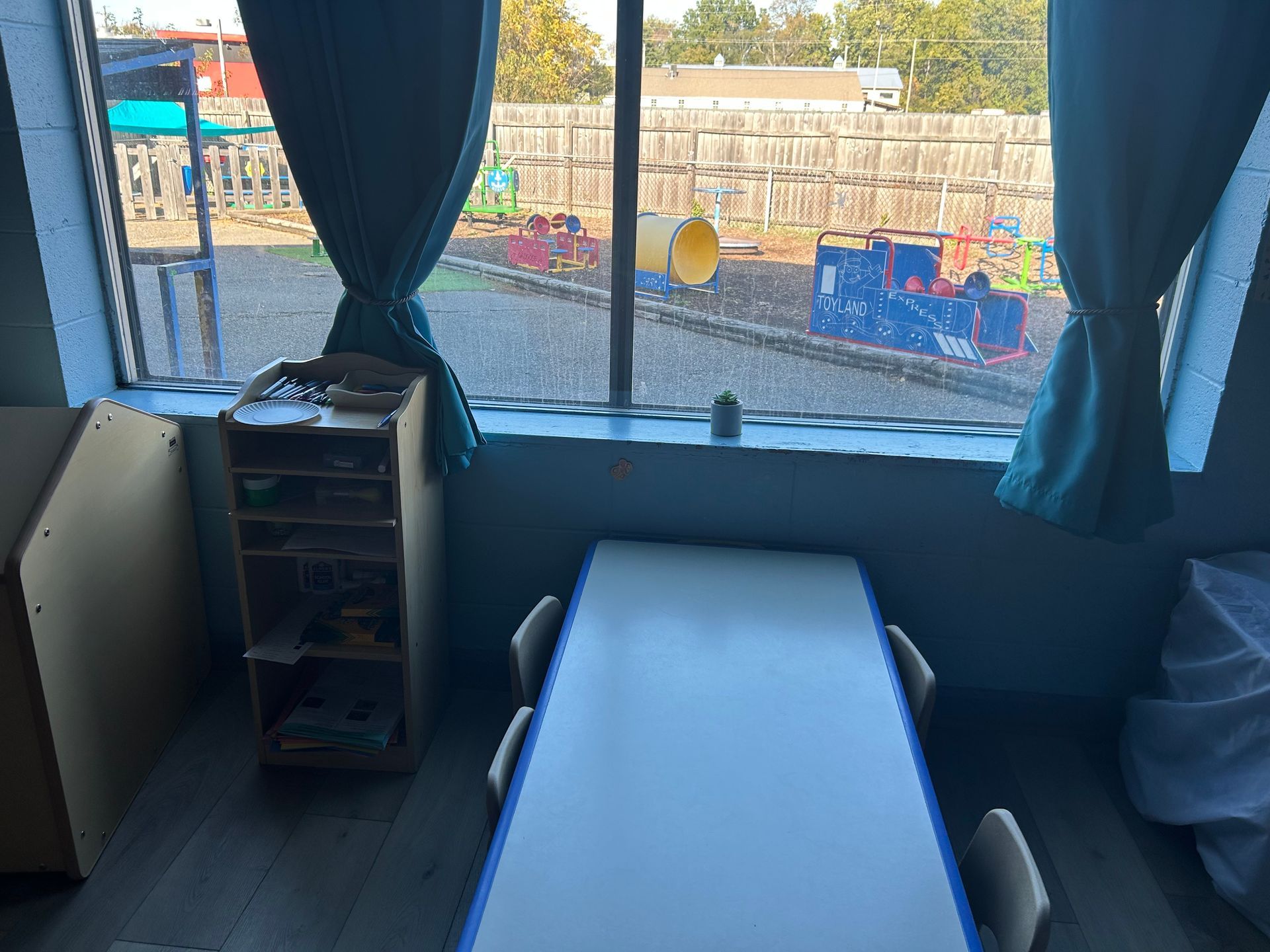 Indoor classroom with a table, chairs, window with curtains, and a view of a playground.