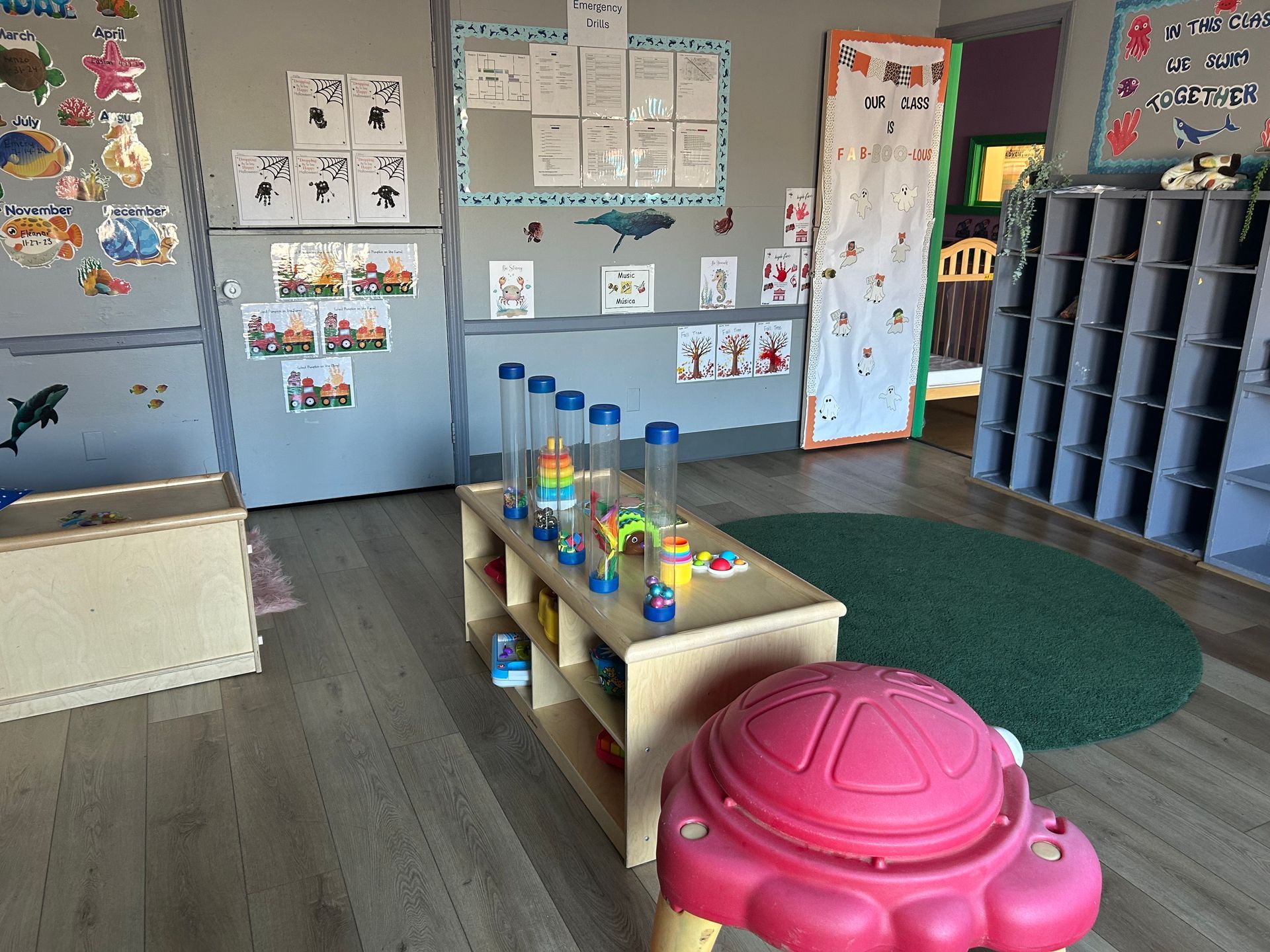 A brightly lit classroom with toy shelves, posters, and a green rug. A red turtle-shaped table is in the foreground.