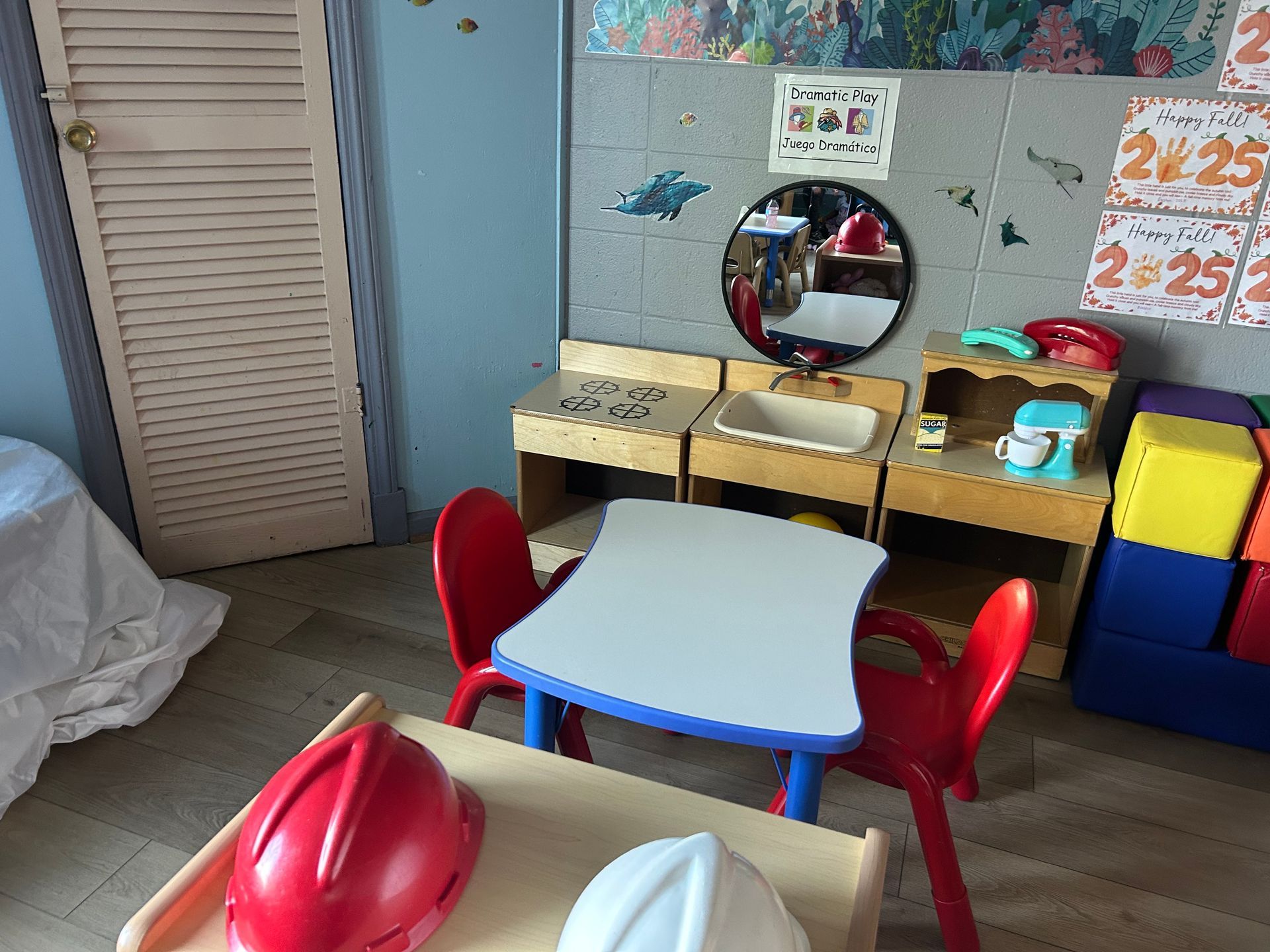Child-sized play area with table, chairs, toy kitchen, and sink; red and white hard hats on a small table.
